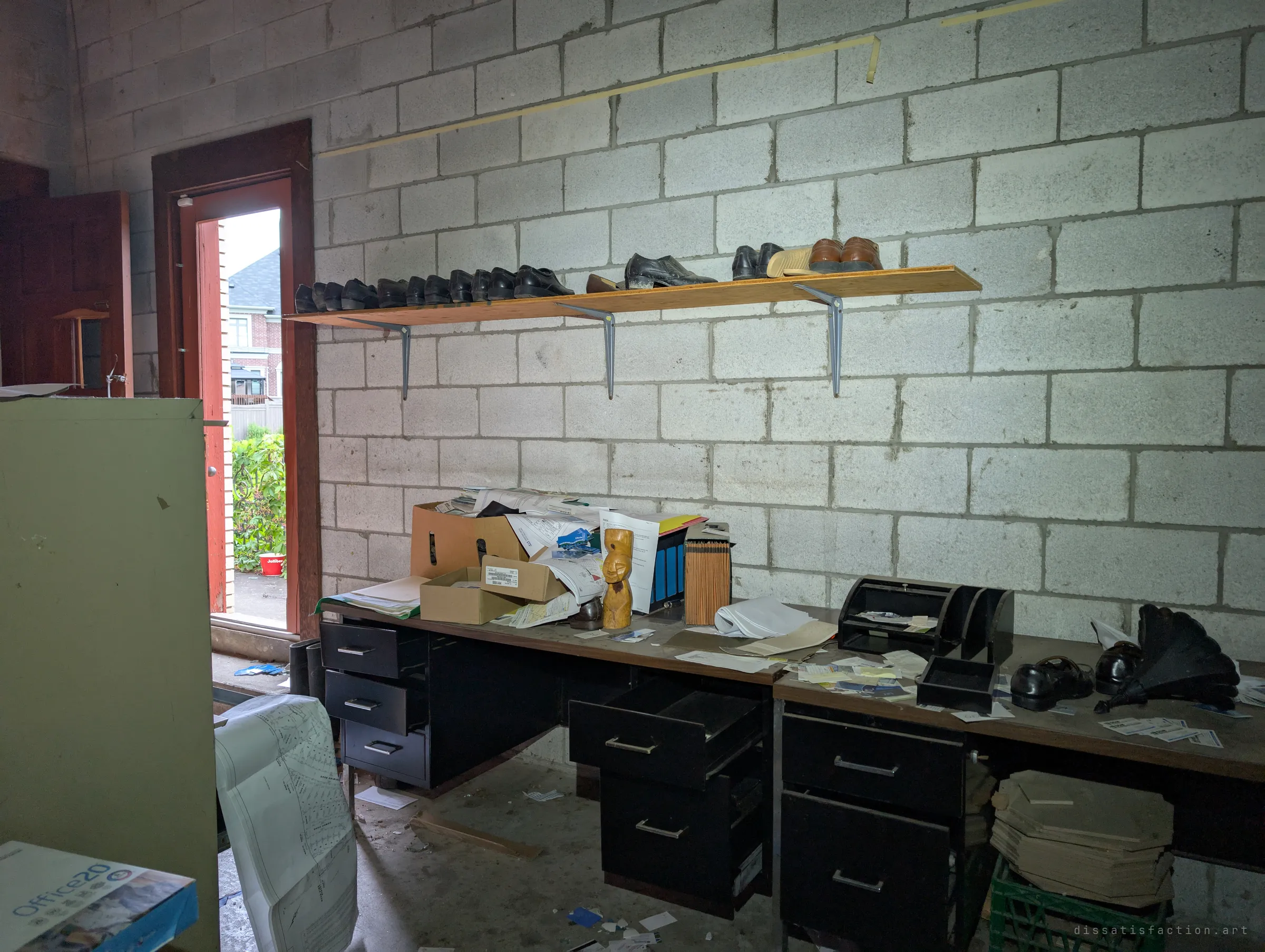 A garage office with a row of dress shoes lined up on a shelf above two cluttered desks of papers, boxes and a carved wooden figure. The open door lets in grey daylight.