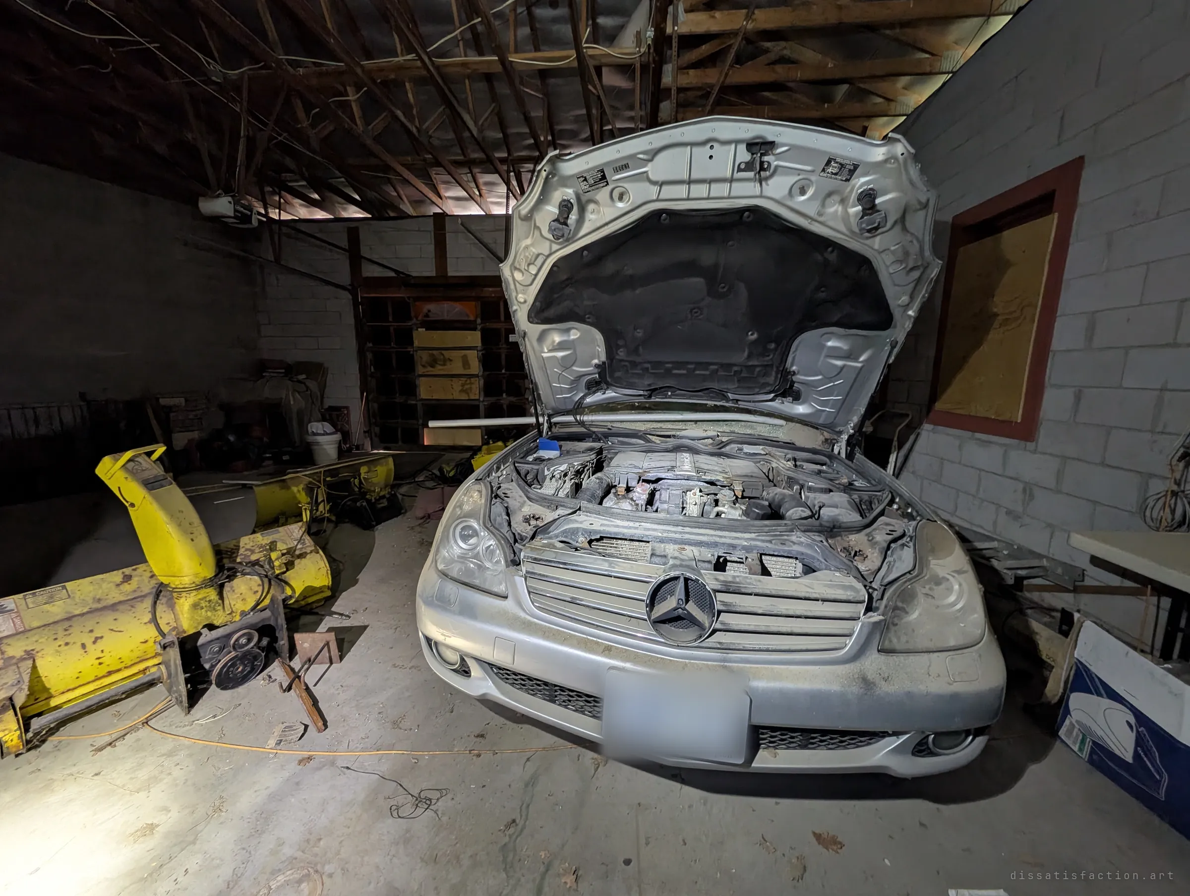 A dusty silver sedan sits in the dark garage with its hood propped open and engine exposed, beside a rusted yellow snowblower attachment. It looks as if someone stopped mid-repair and never came back.