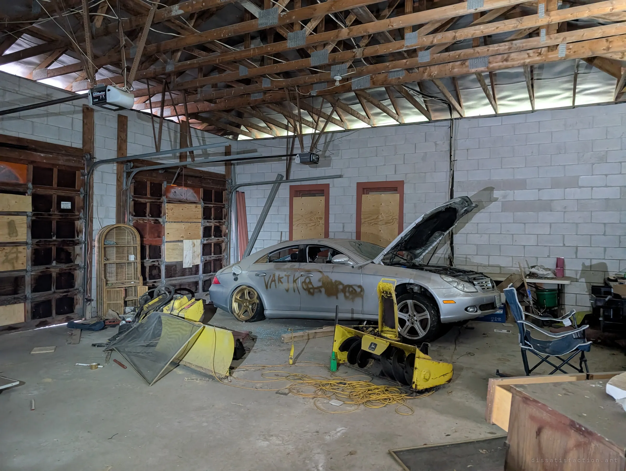 The wide garage under exposed roof trusses, with the tagged silver sedan on a flat gold wheel, boarded windows and doors, and yellow snowblower parts tangled in extension cord. The whole space feels frozen in place.