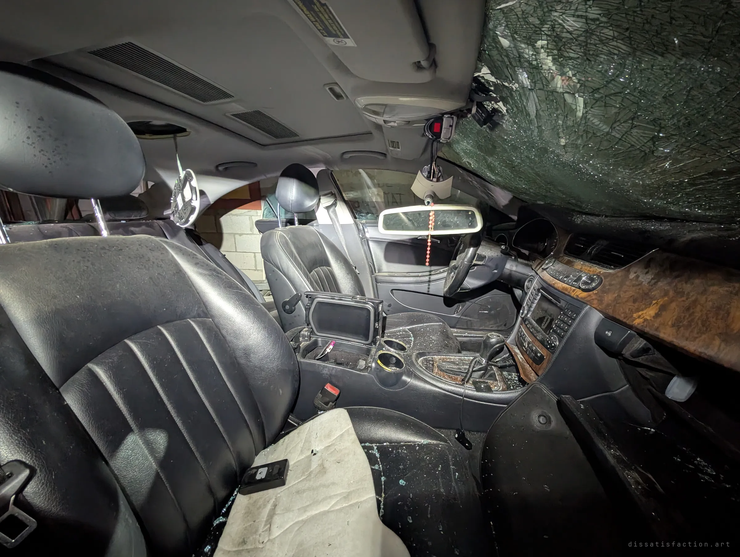 Inside the abandoned sedan, black leather seats are strewn with safety glass under a caved-in, spiderwebbed windshield, and a bead chain still hangs from the mirror.