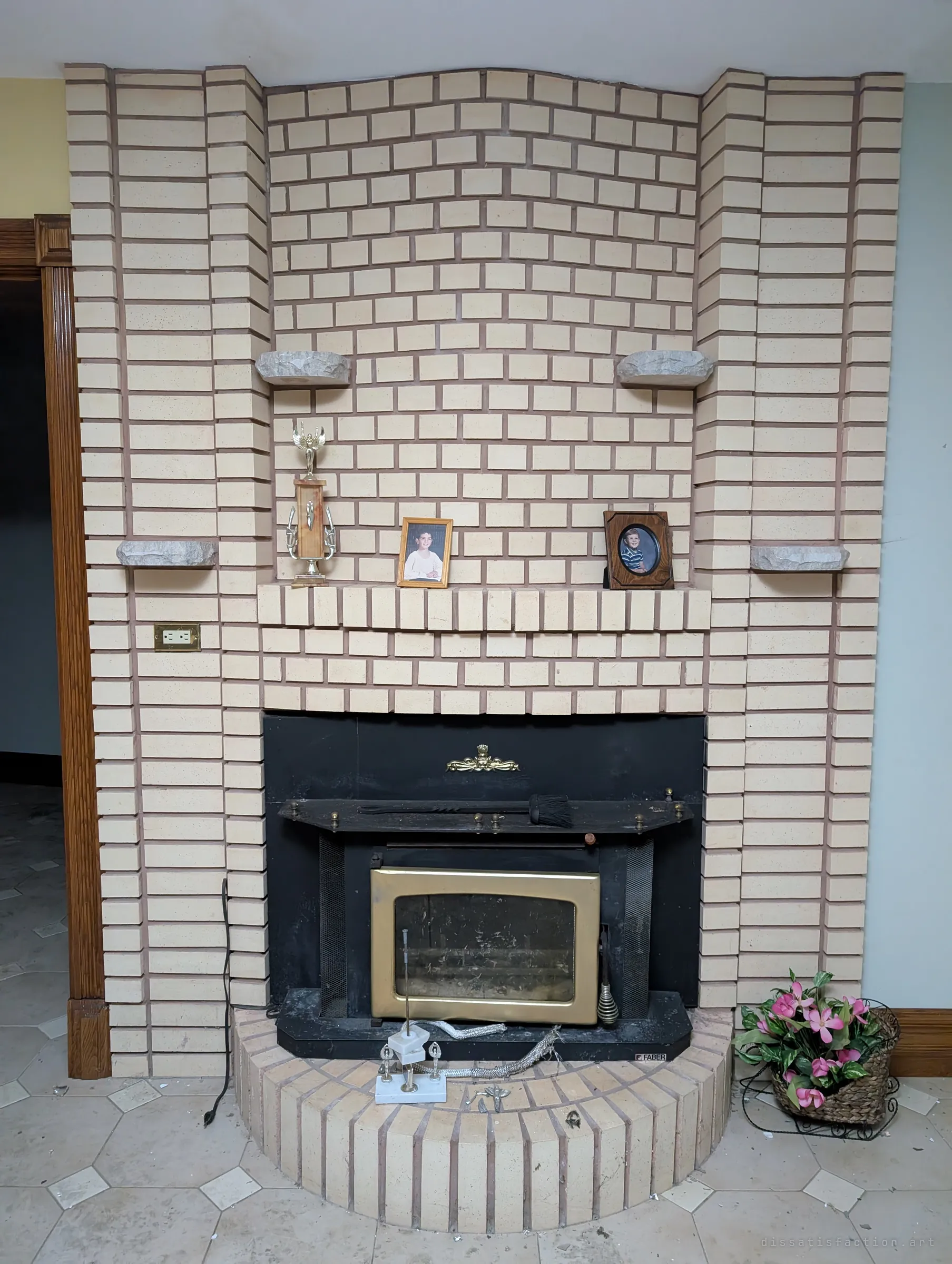 A curved cream-brick fireplace with a brass-trimmed insert, a trophy and framed school portraits still on the mantel. The family photos left in place show how suddenly the house was abandoned.