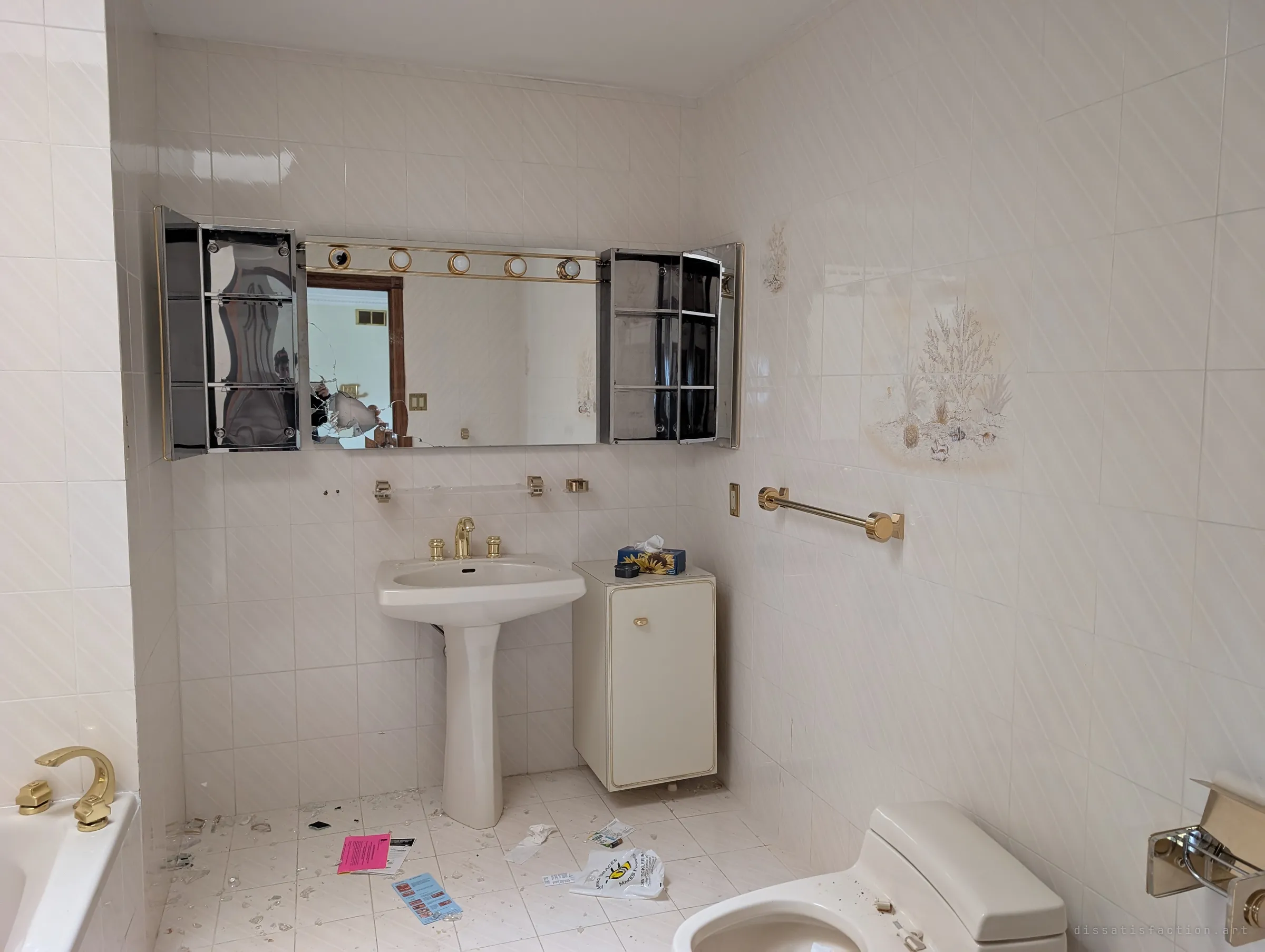 A cream-tiled bathroom with gold fixtures, open mirrored cabinets and a smashed mirror, glass shards and papers scattered across the floor.
