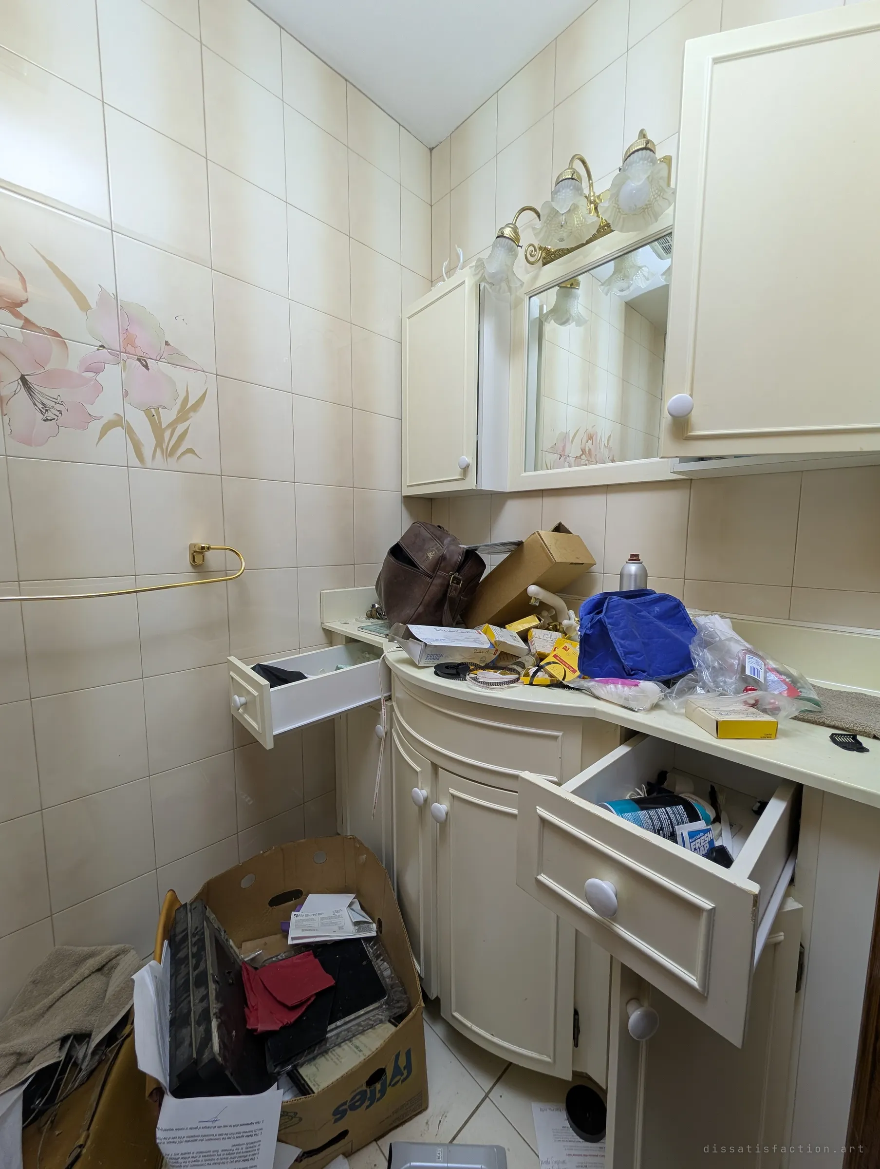 A small powder room with floral painted tiles, open drawers and a vanity heaped with bags, boxes and toiletries, a cardboard box of papers on the floor. The clutter shows the house was left with belongings still inside.