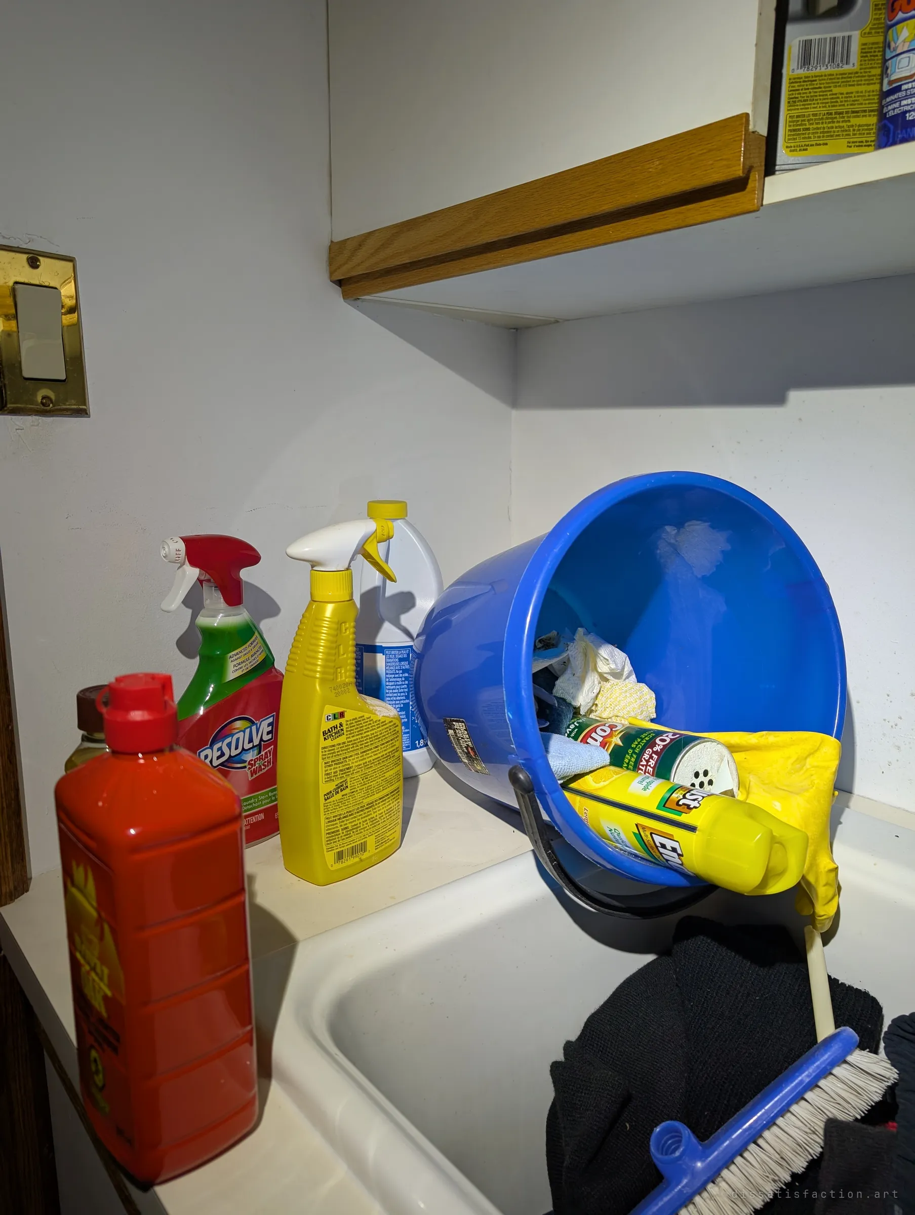 Cleaning sprays, a bottle of floor soap and a tipped blue bucket with gloves and a scrub brush on a laundry sink, lit by a harsh flashlight beam.