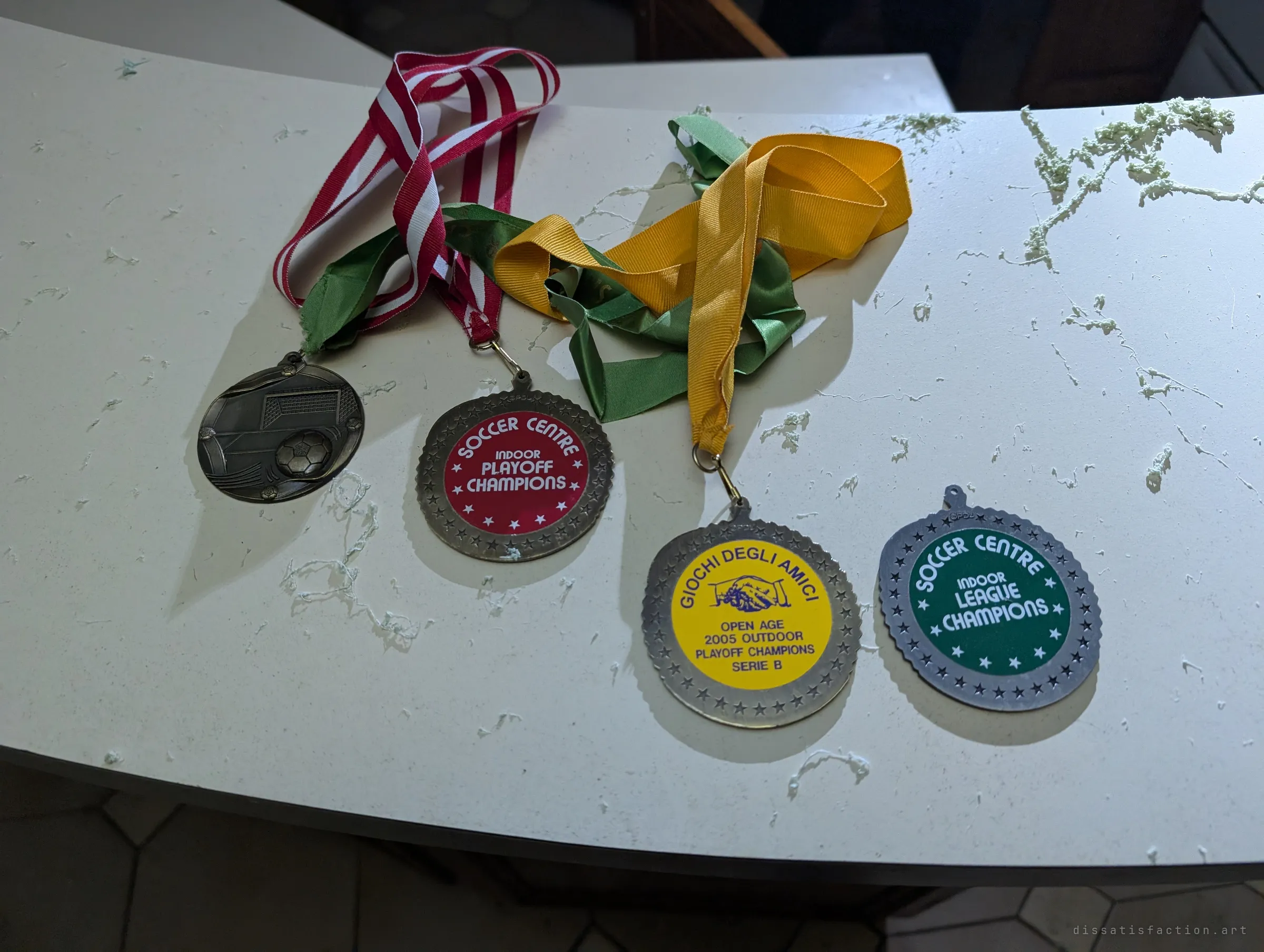 Four soccer medals with faded ribbons lie on a white counter dusted with debris, leftover trophies of someone's playing years.