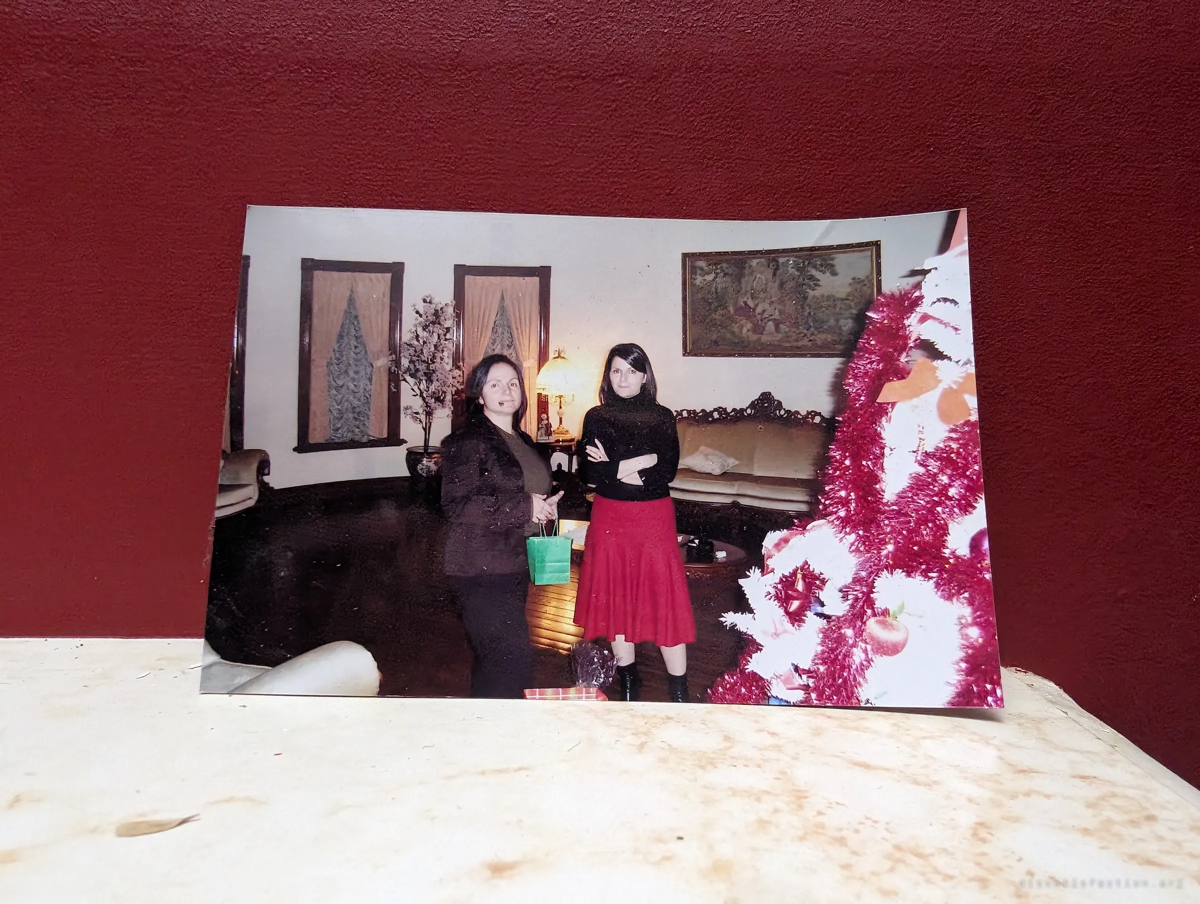 An old printed snapshot propped against a deep red wall shows two women standing in an ornate living room beside a pink-tinselled Christmas tree, a glimpse of the house when it was lived in.