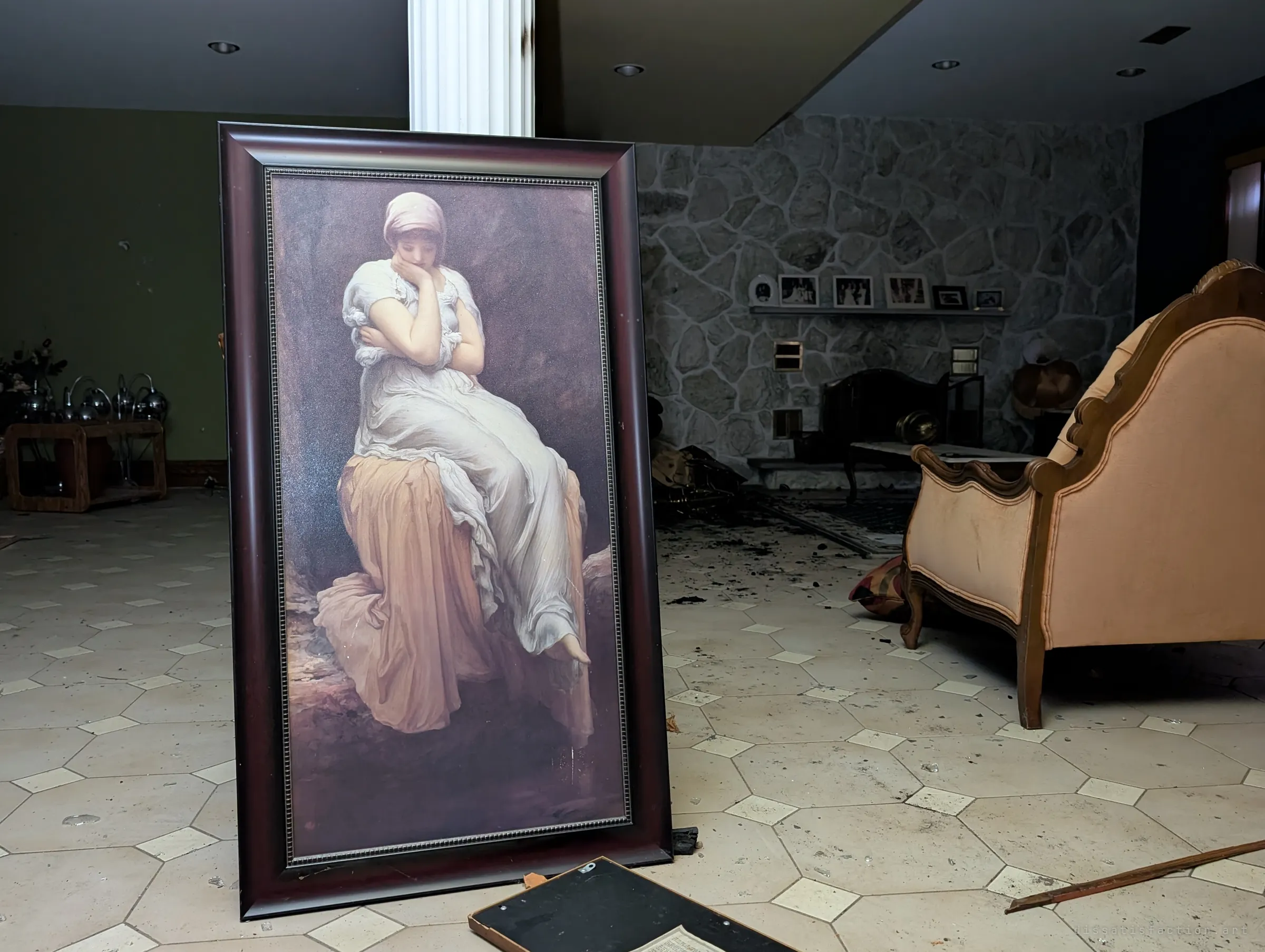 A large framed print of a pensive woman in a white gown leans on the tiled floor of a dim living room, a peach armchair and a stone fireplace wall with family photos behind it.