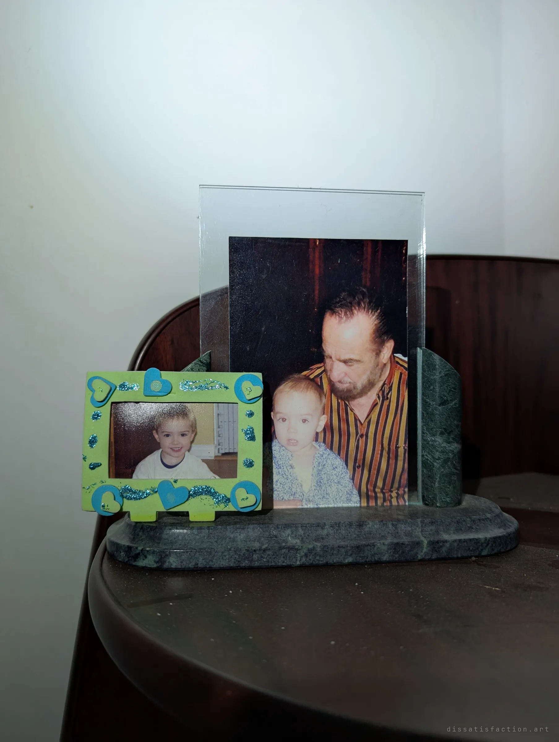 A marble photo stand on a dark wooden table holds a picture of an older man with a toddler and a small craft frame of a smiling child.