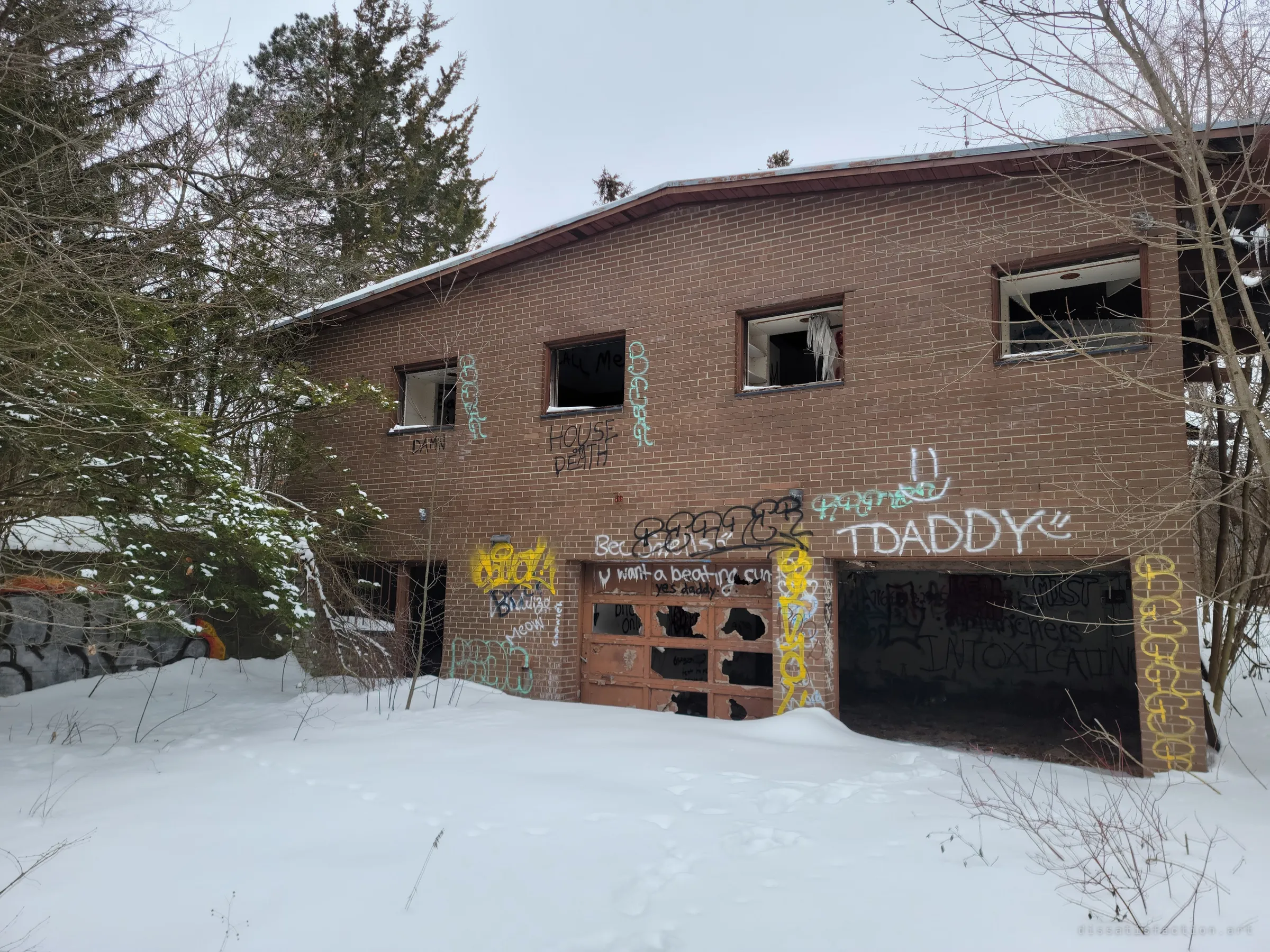 A two-storey brick house sits half-buried in snow among pines, its windows smashed and its garage doors covered in graffiti. The establishing shot of the whole abandoned building.