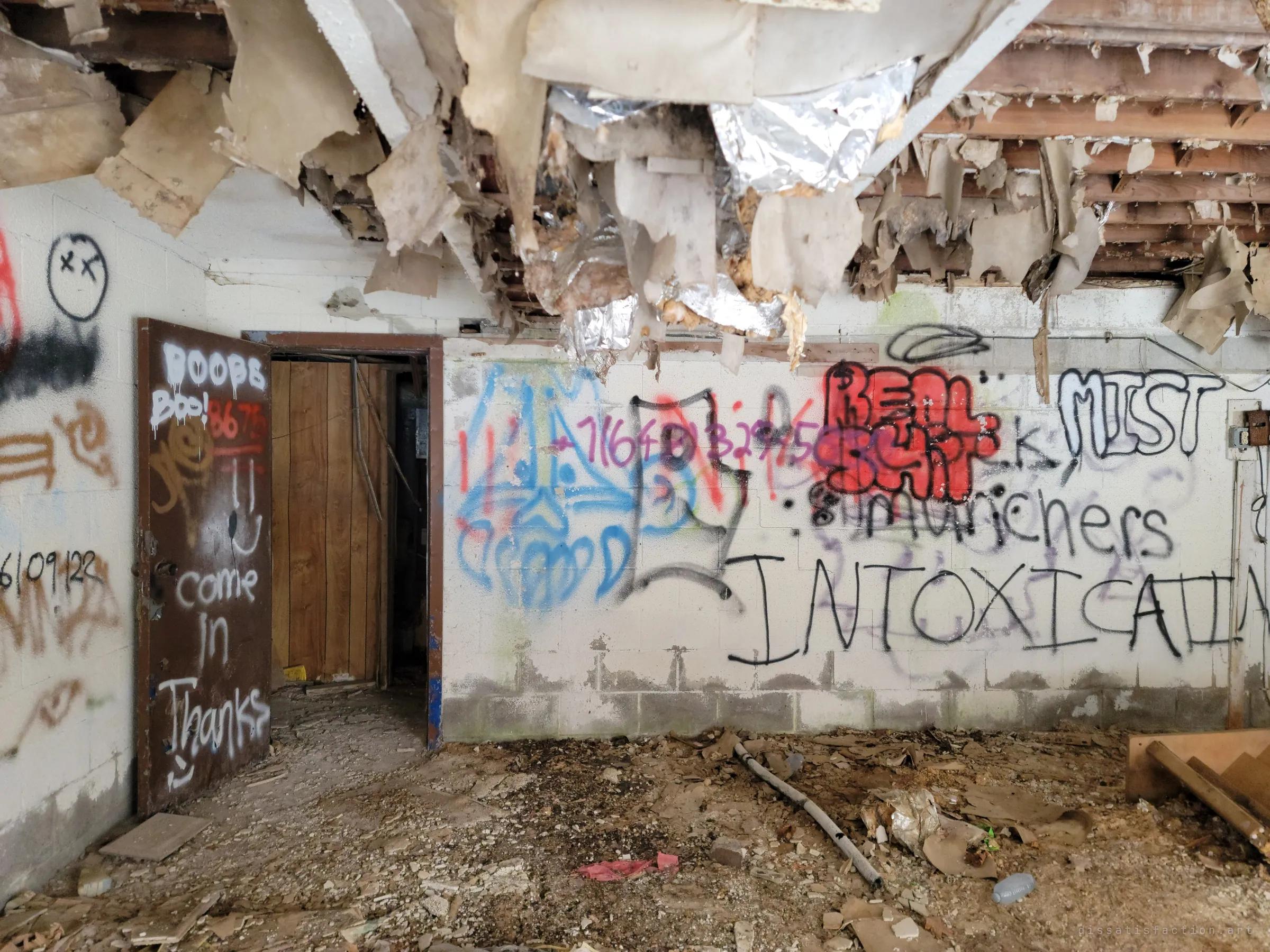 Inside the garage, the ceiling has collapsed into shreds of insulation and foil above a graffiti-covered block wall and an open doorway. Debris covers the floor.