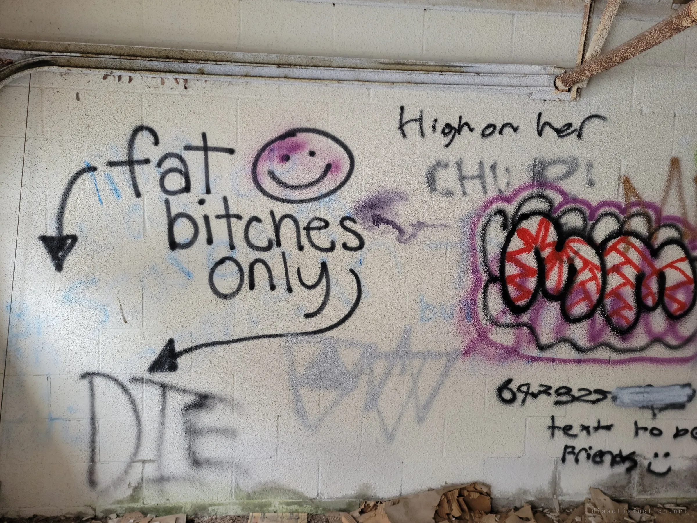 A close-up of a painted block wall crowded with crude graffiti slogans, a smiley face and a large bubble tag. It shows how thoroughly visitors have written over the place.