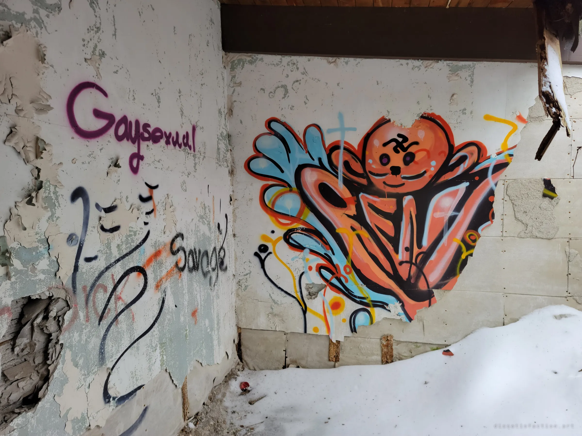 A colourful orange and blue graffiti character spreads across a peeling wall in a snow-drifted corner, beside tags on a curved wall. Snow has blown in under the roof.