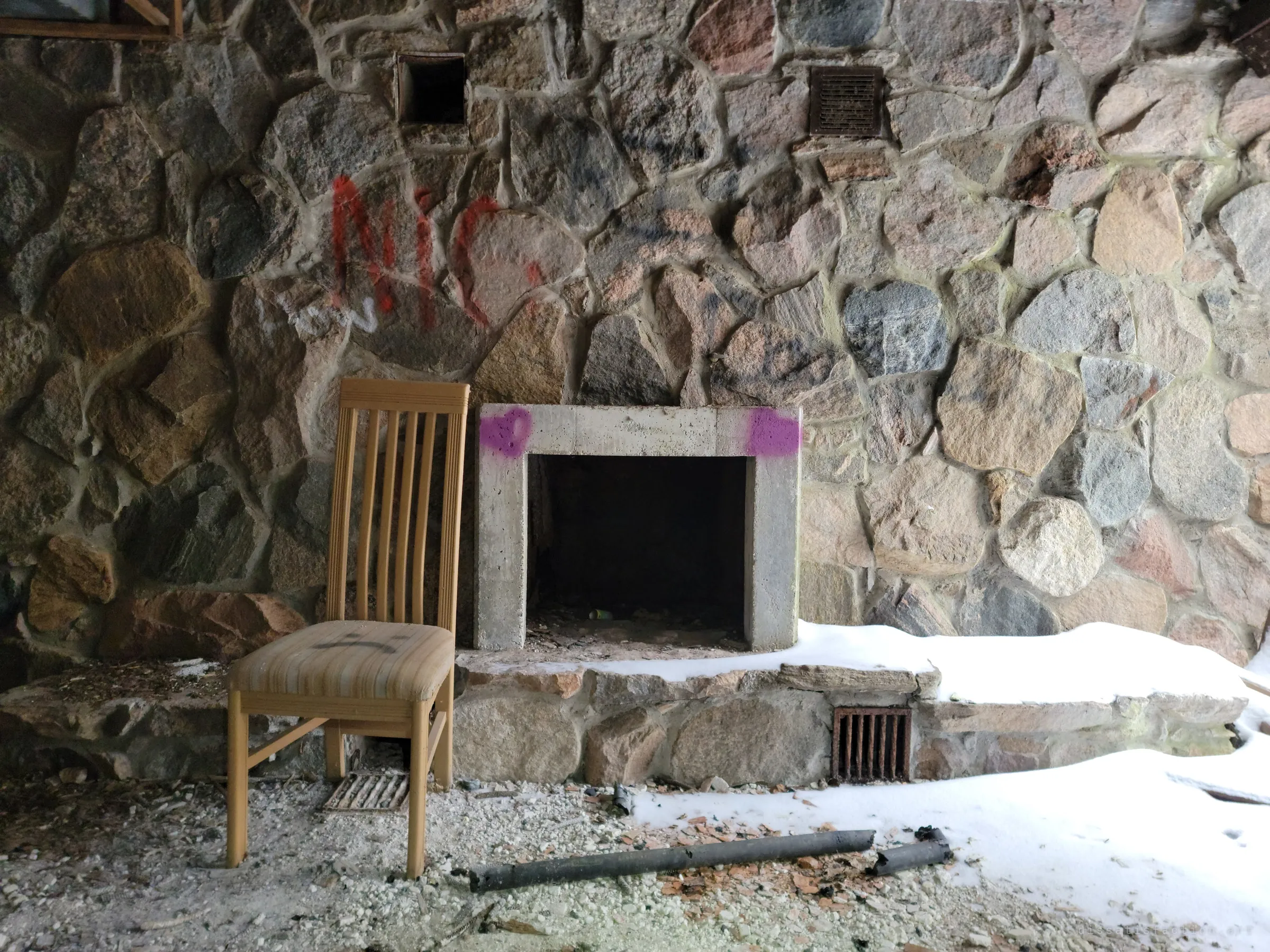 A wooden dining chair stands alone beside a concrete-framed fireplace set into a fieldstone wall, with snow drifted across the hearth. A quiet domestic leftover in the ruin.