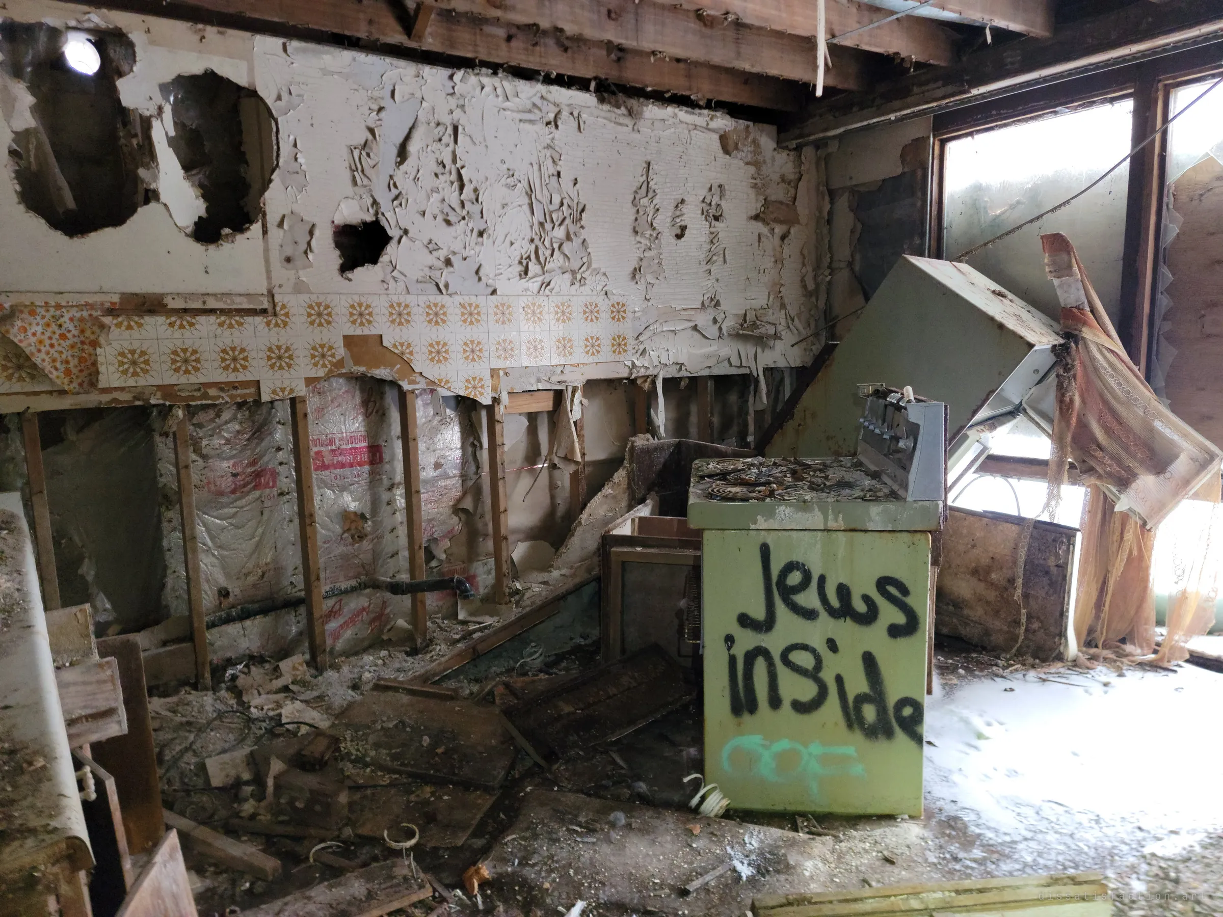 A gutted kitchen with walls stripped to the studs, patterned tile clinging on and a toppled range hood hanging over a graffitied stove. Snow and light pour in through broken windows.
