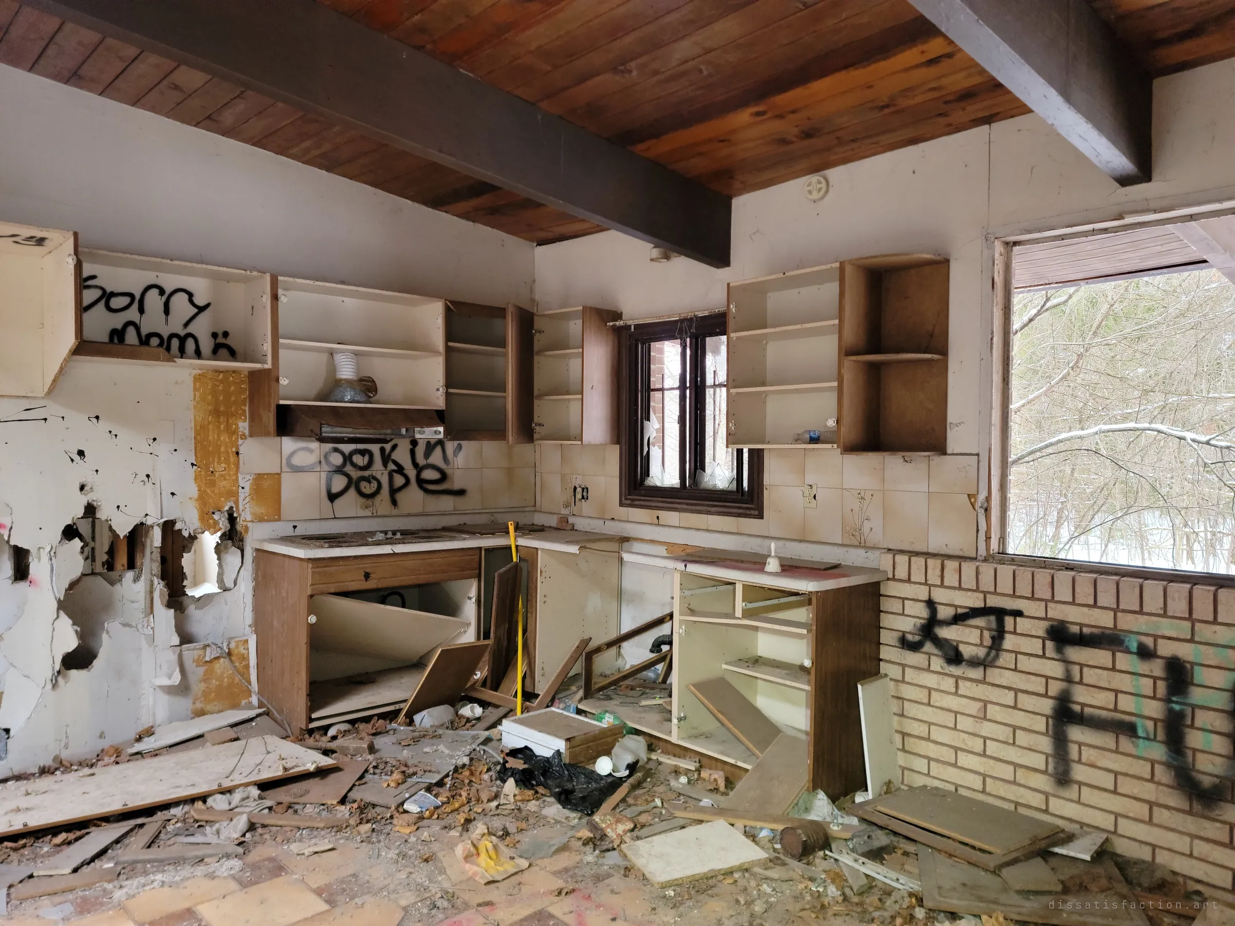 A gutted kitchen under a dark-beamed wood ceiling, cabinets hanging open and tagged with spray paint, the floor buried in broken doors and debris. A glassless window frames snowy bare woods.