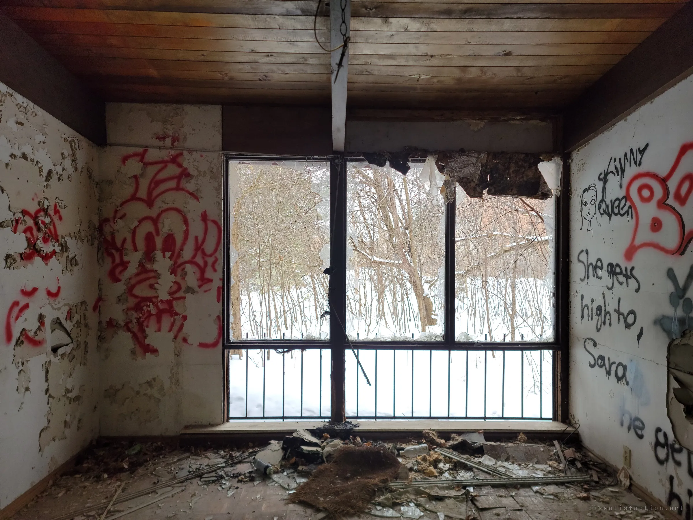 A bare room with peeling walls covered in red and black graffiti, facing a large shattered window with a railing and snow-covered woods beyond. Scorched ceiling material hangs over the glass.