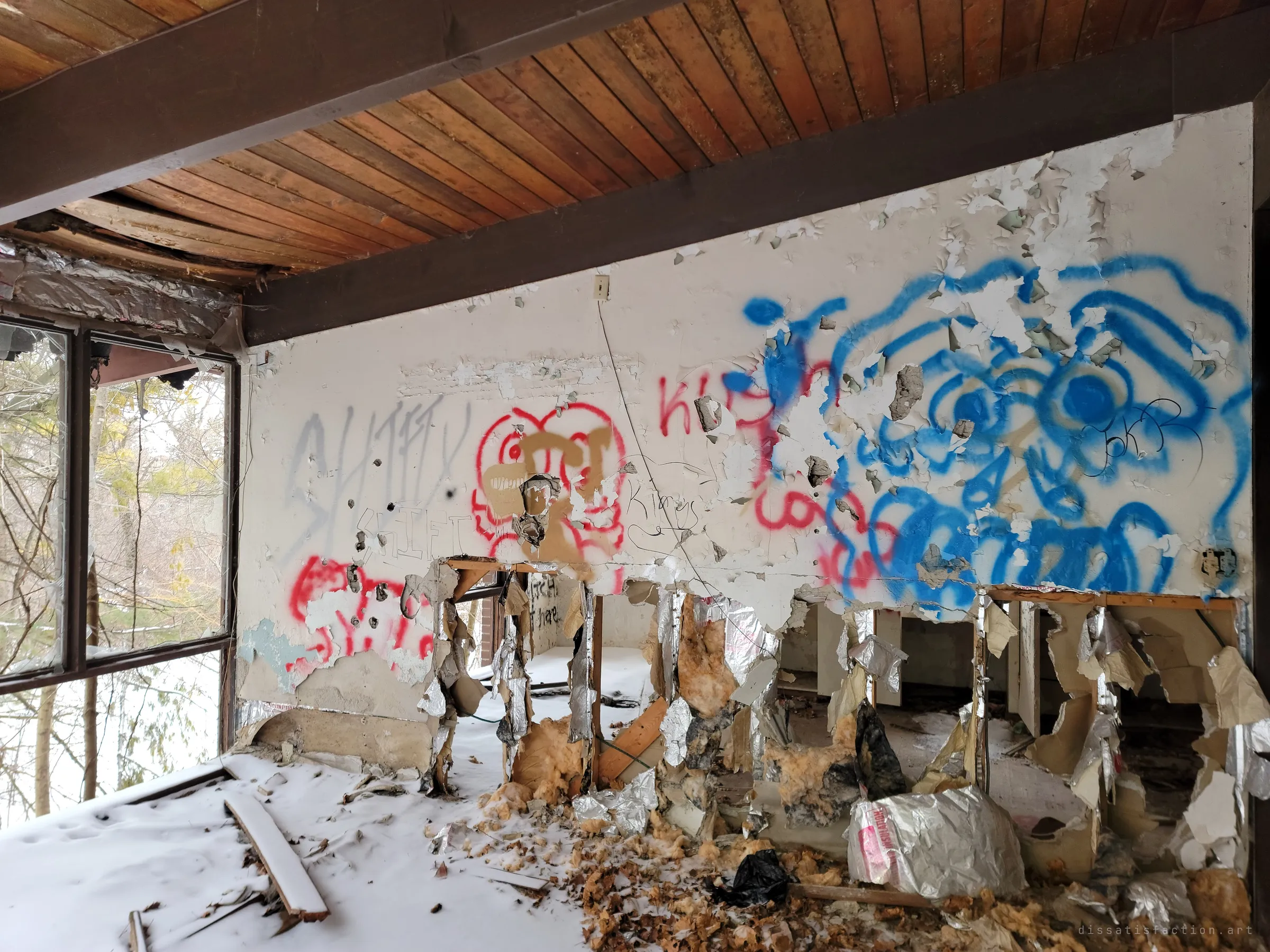A wall of blue and red graffiti ripped open along its base, spilling foil-backed insulation onto a snow-dusted floor. Broken corner windows look out onto trees.