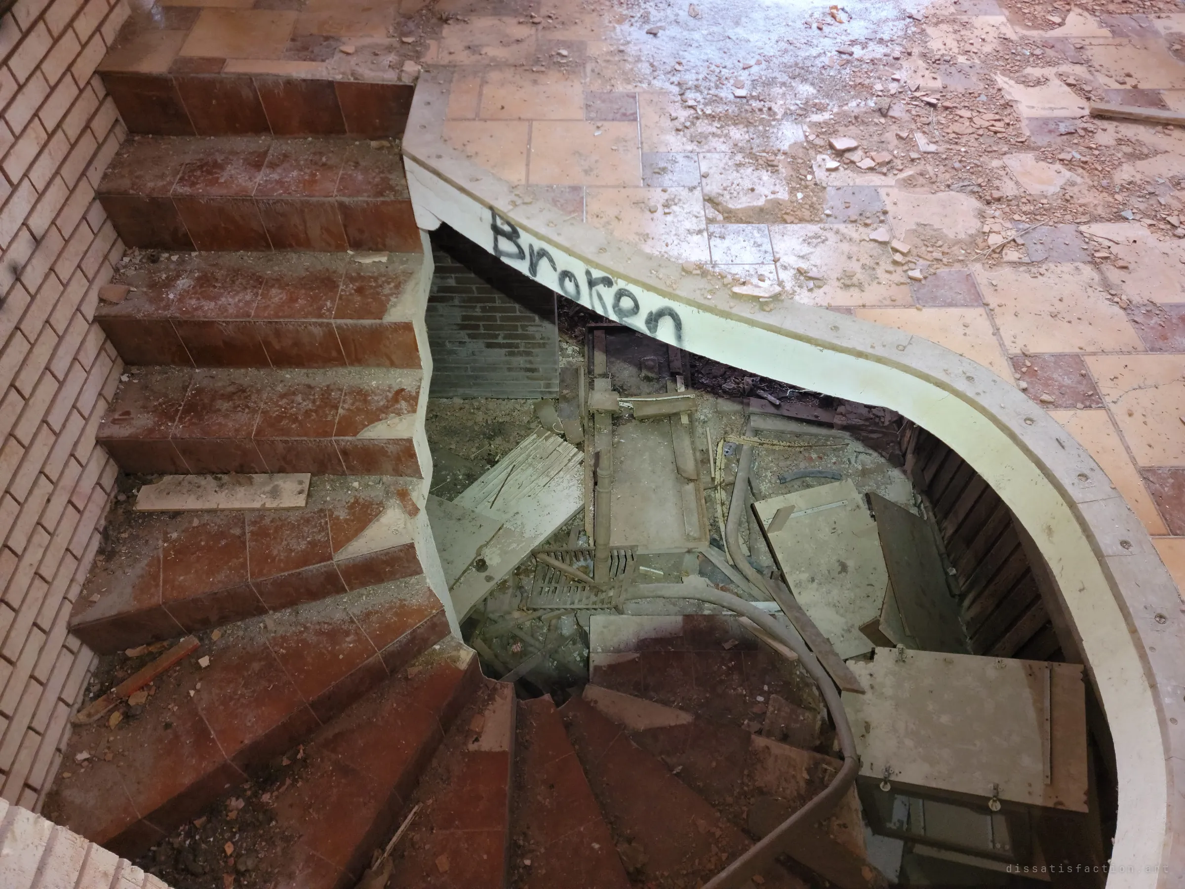 Looking down a curved tiled staircase whose lower flight has collapsed into a pit of wreckage, a single word sprayed on its edge. The tiled landing above is littered with shards.