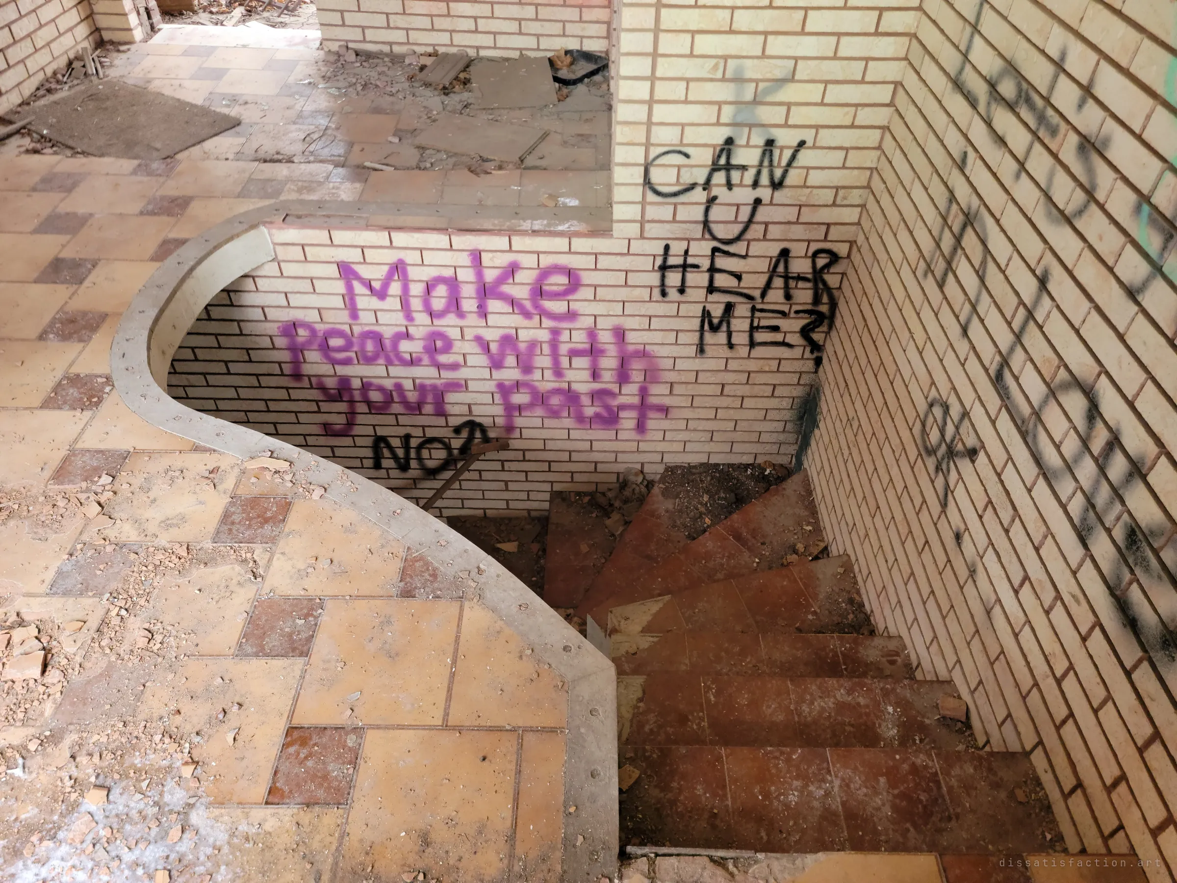 A spiral stair winds down beside a curved tiled landing, the cream brick walls sprayed with purple and black messages about the past. Debris and loose tiles cover the floor.
