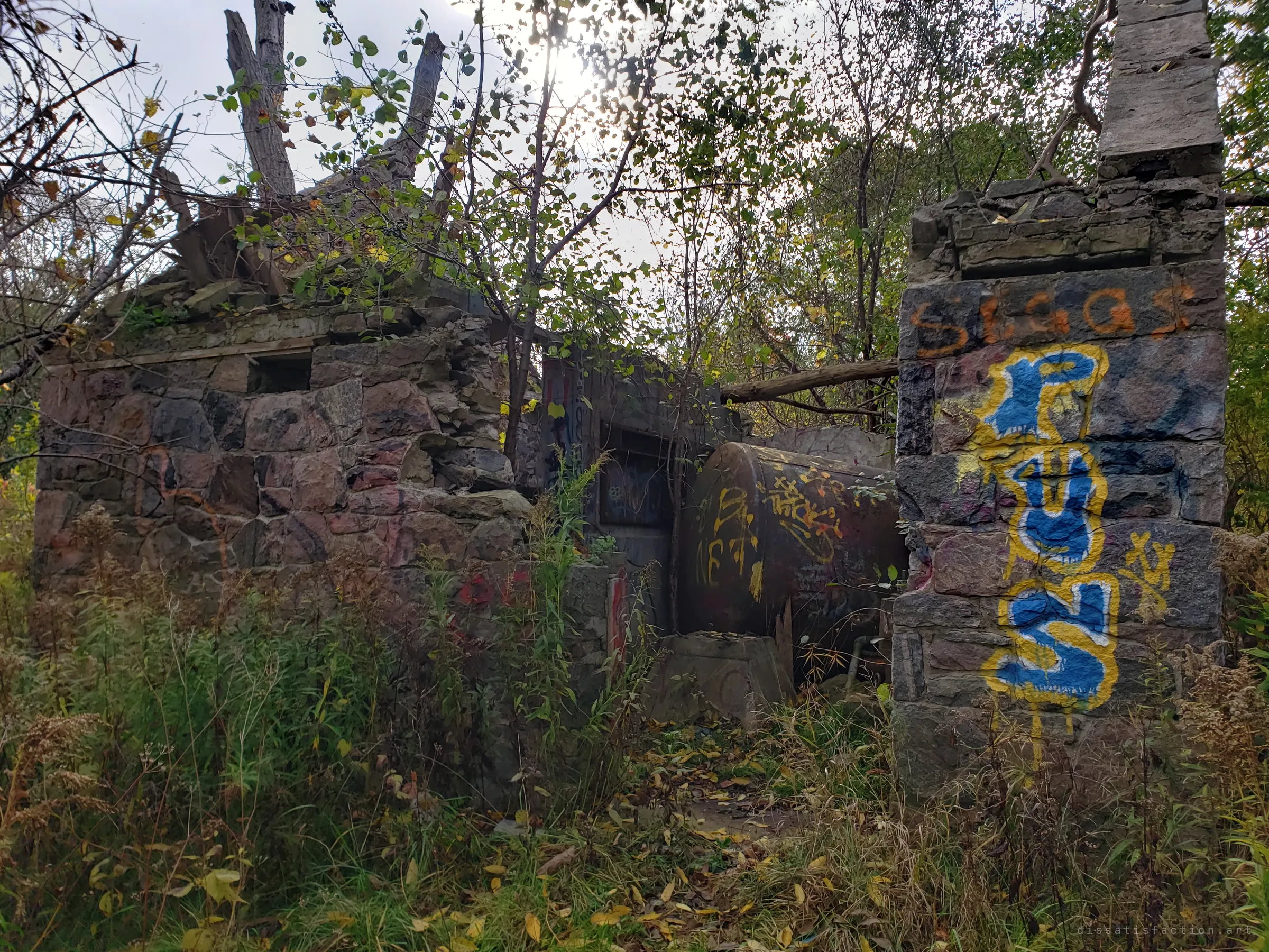 Collapsed fieldstone walls and a toppled steel tank sit in tall weeds, with a heavily tagged stone pillar in the foreground. The roof is gone and trees now grow through the ruin.