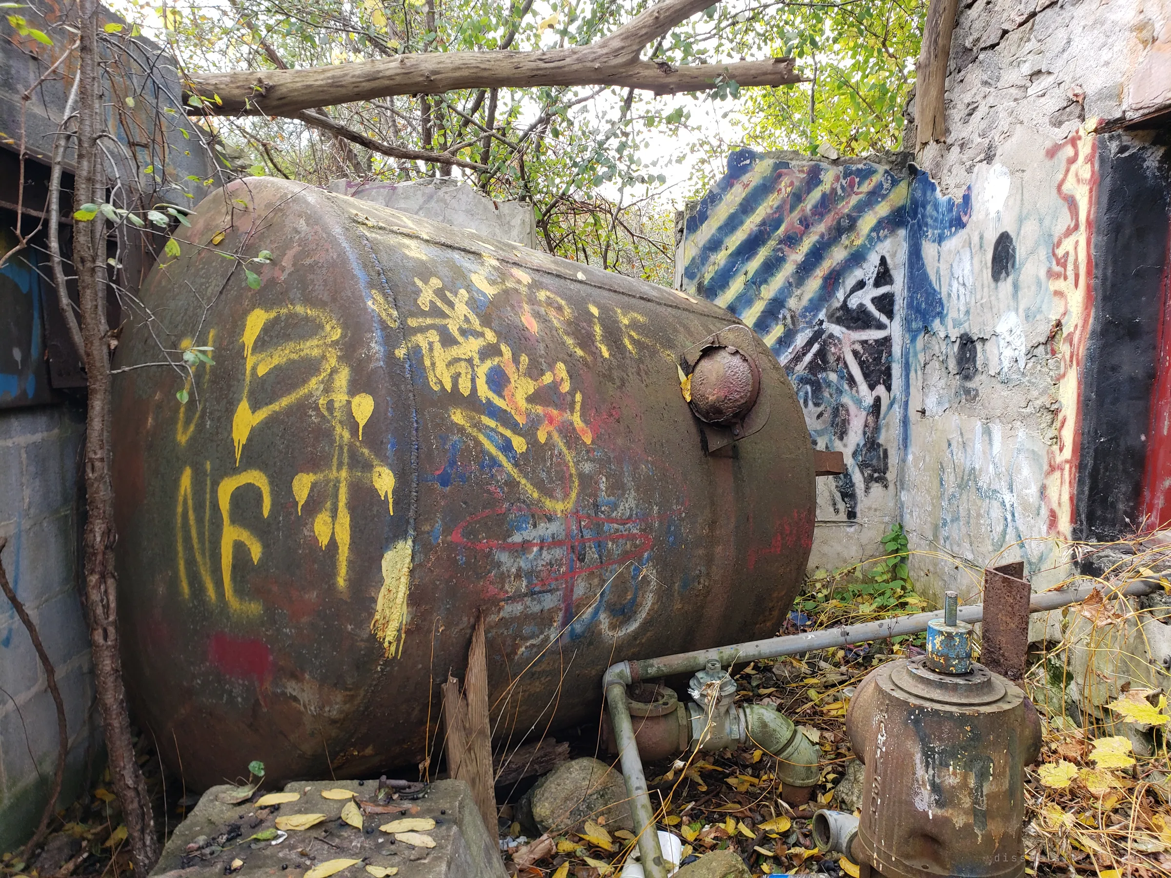A large rusted pressure tank covered in spray paint lies inside the roofless pumphouse, beside old piping and a pump housing among fallen leaves. It is the main piece of machinery left behind.