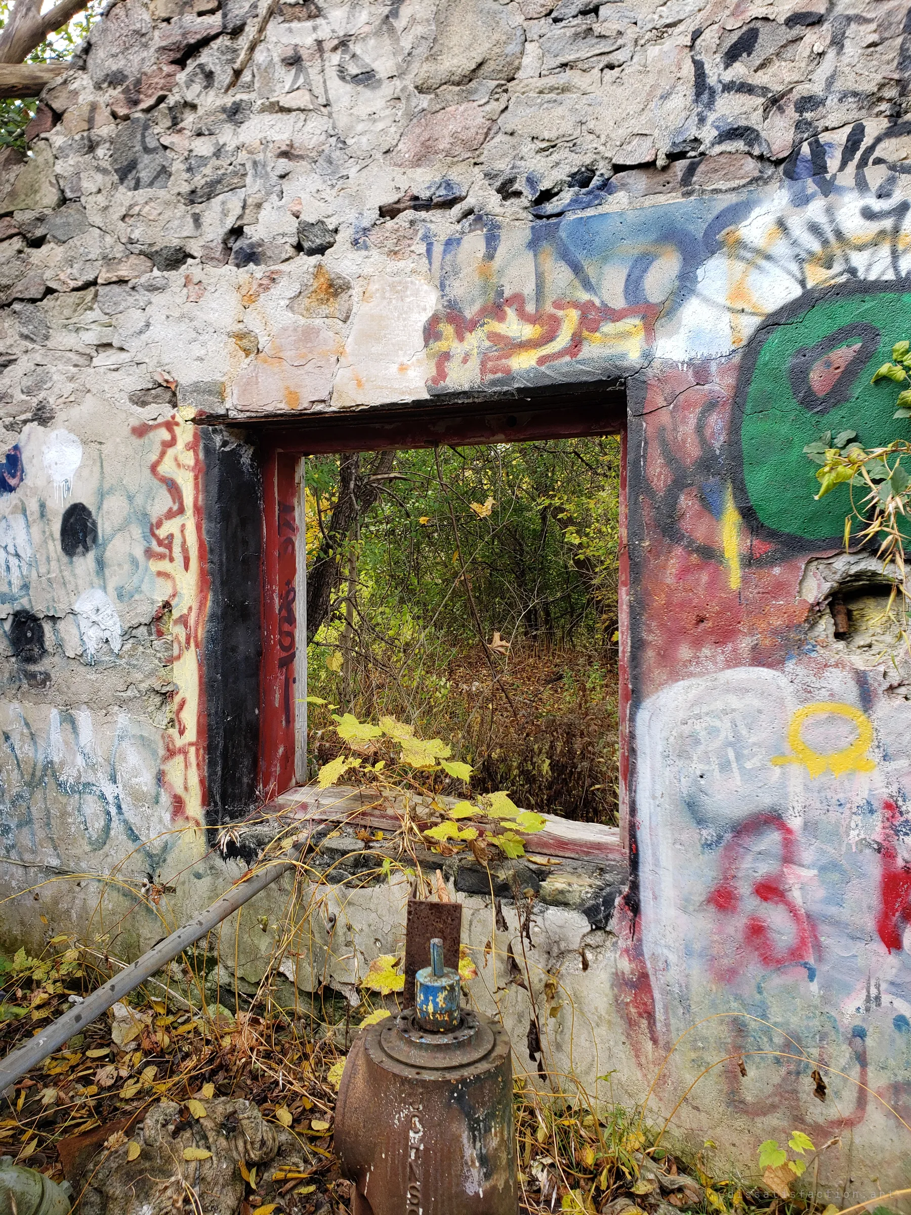 A red-framed window in a graffiti-covered stone wall looks out onto dense autumn woods, with a rusted pump head and pipe in the foreground. It frames how the forest has closed in around the building.