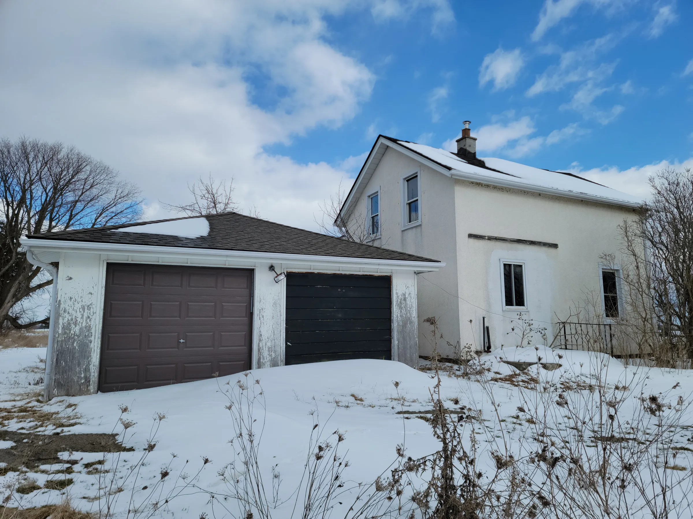 A peeling white two-car garage with mismatched brown and black doors sits beside the stucco house, snow drifted against both. Small repairs like the newer shingles and door show someone kept it up not long ago.