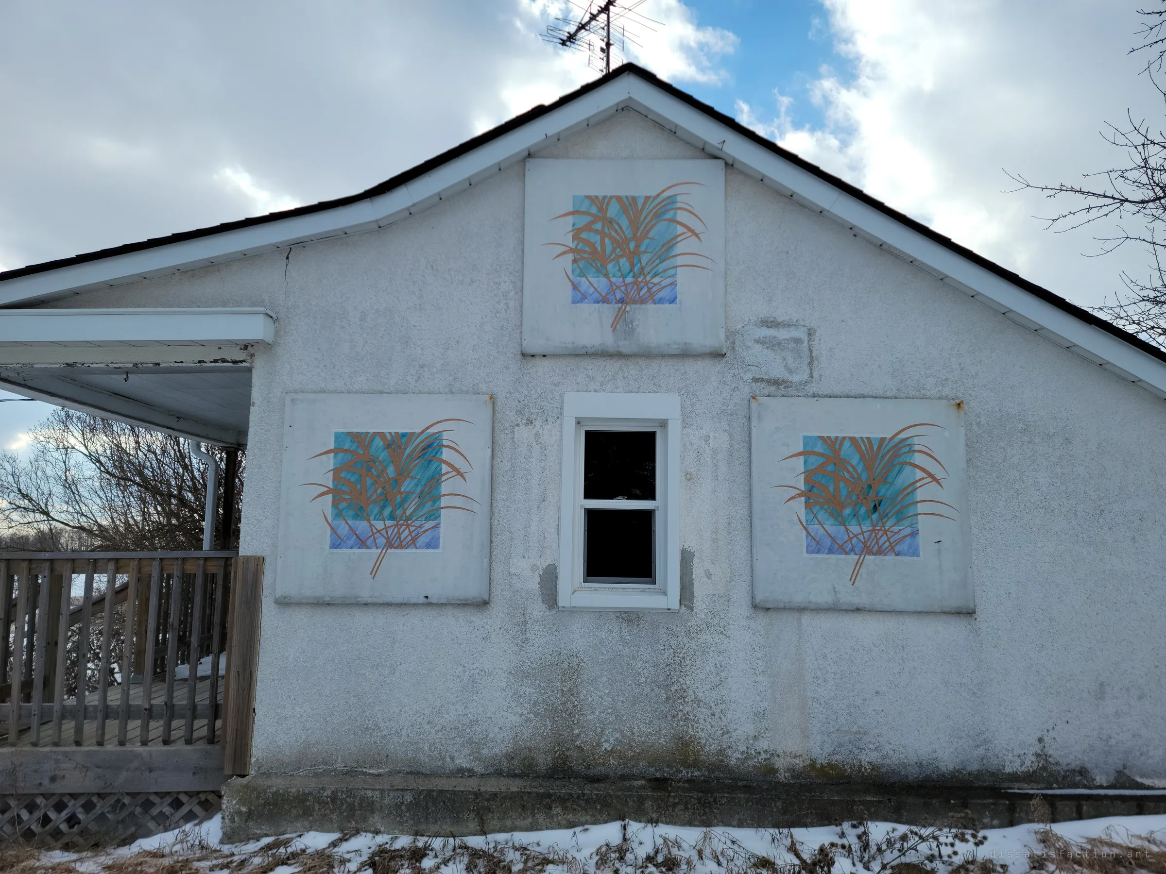 The gable end of the house, its grey stucco decorated with three painted panels of copper grasses over blue squares around a single small window. A homely, hand-made touch left behind on an otherwise stripped building.