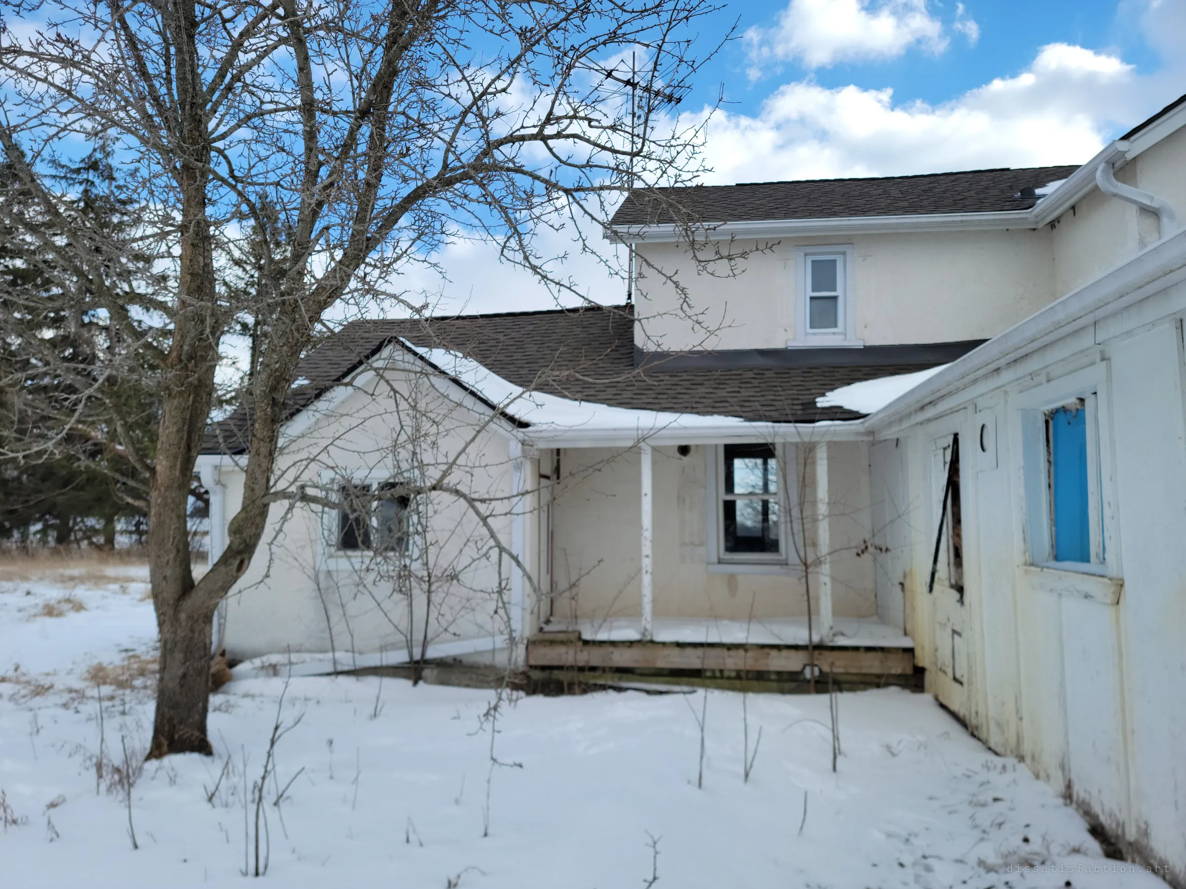 The back of the house behind a bare tree, with a small snow-covered porch and a side addition whose window is boarded with bright blue panel. Snow lies undisturbed right up to the step.