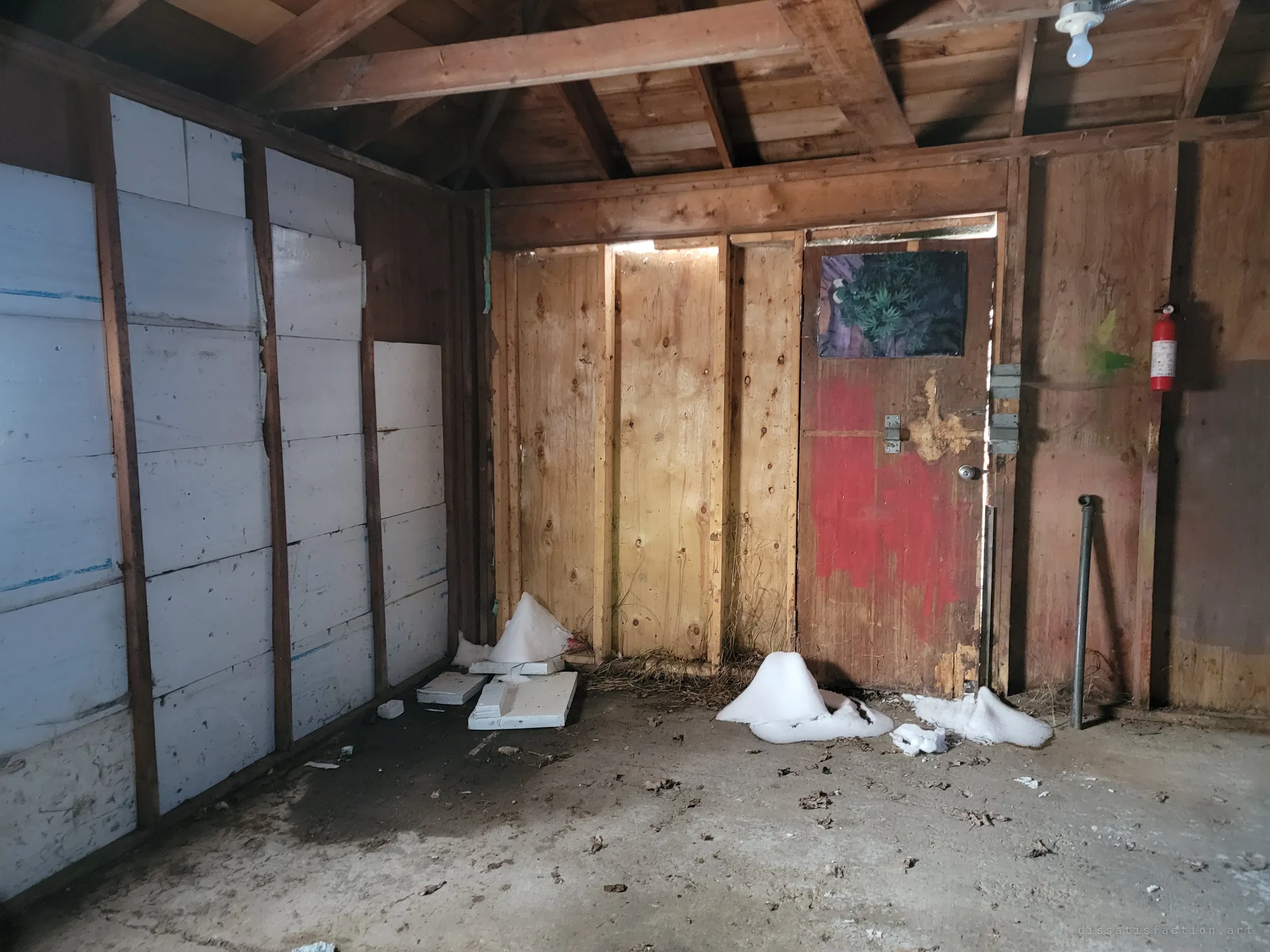 Inside the garage: raw wood walls lined with foam insulation, a red-smeared door with a plant poster, a fire extinguisher and small drifts of snow blown in across the concrete floor.