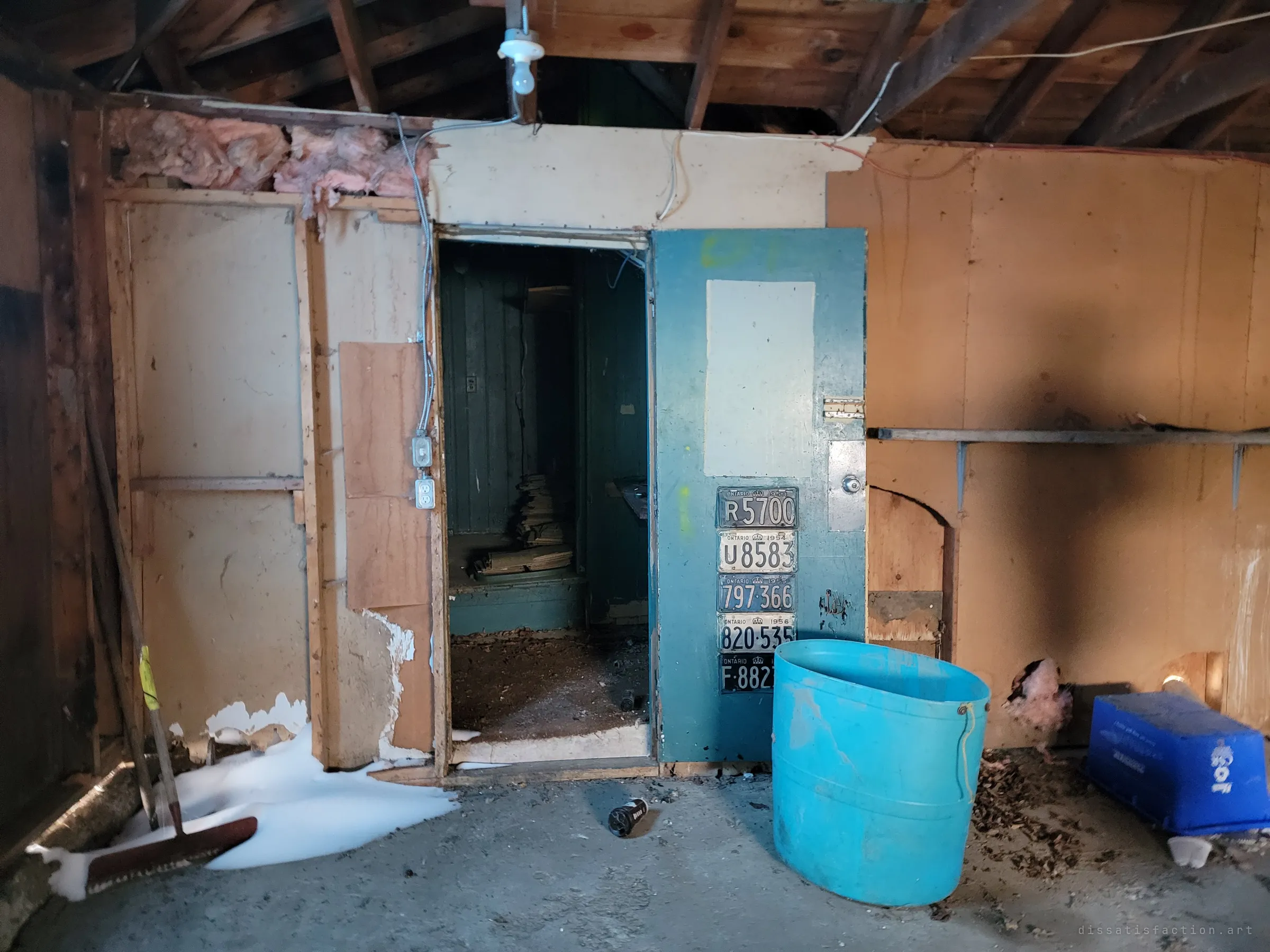 A gutted back room with insulation spilling from the ceiling, an open doorway into a teal closet, and a door hung with a column of old licence plates beside a blue plastic tub.