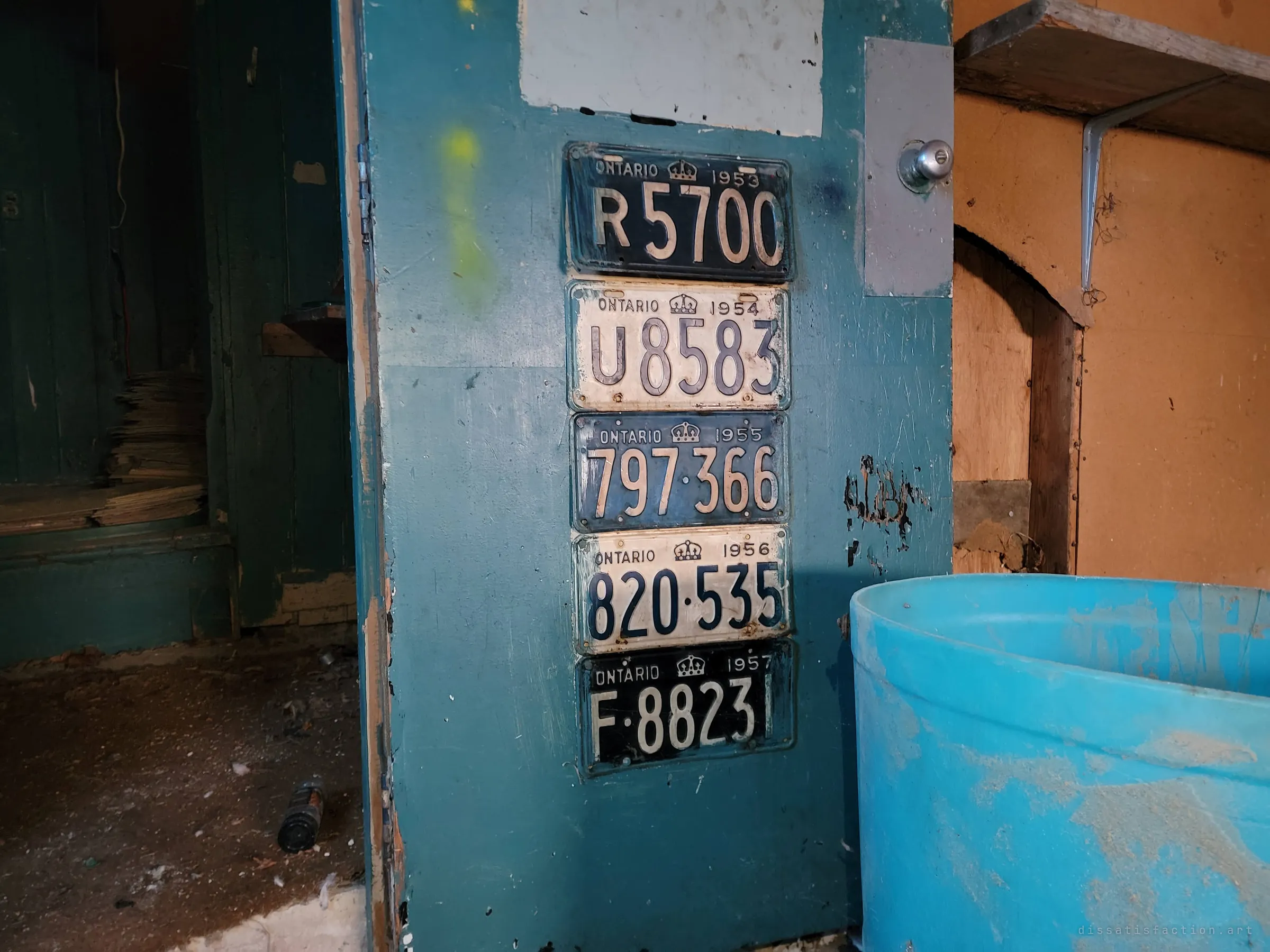 Five old licence plates from the mid-1950s mounted in a column on a scuffed teal door. A collector's keepsake still hanging in a house stripped of almost everything else.