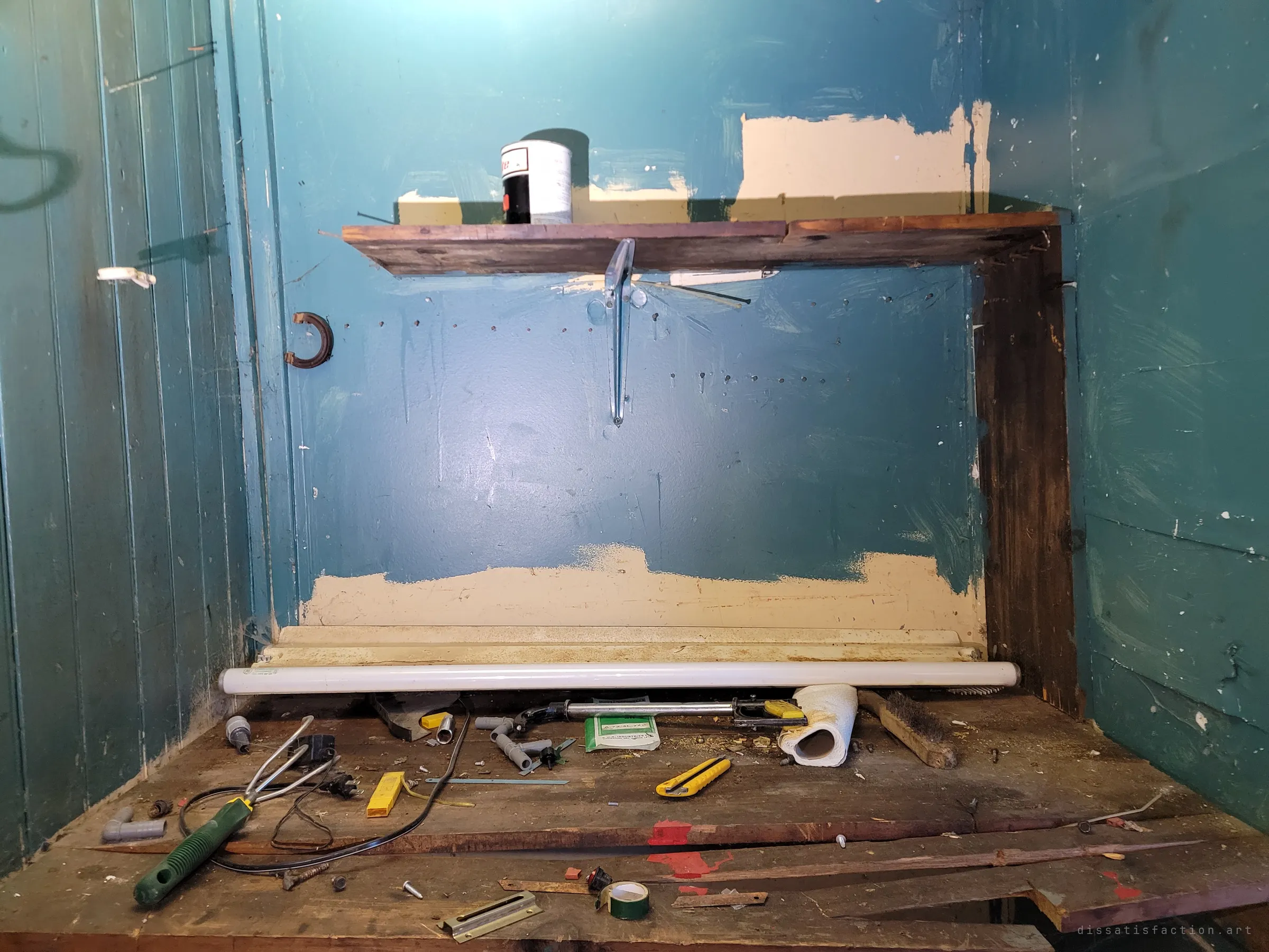 A teal-painted workbench nook littered with tools, pipe fittings, a box cutter and a fluorescent tube, under a single shelf holding a paint can. The last workshop left as it was dropped.