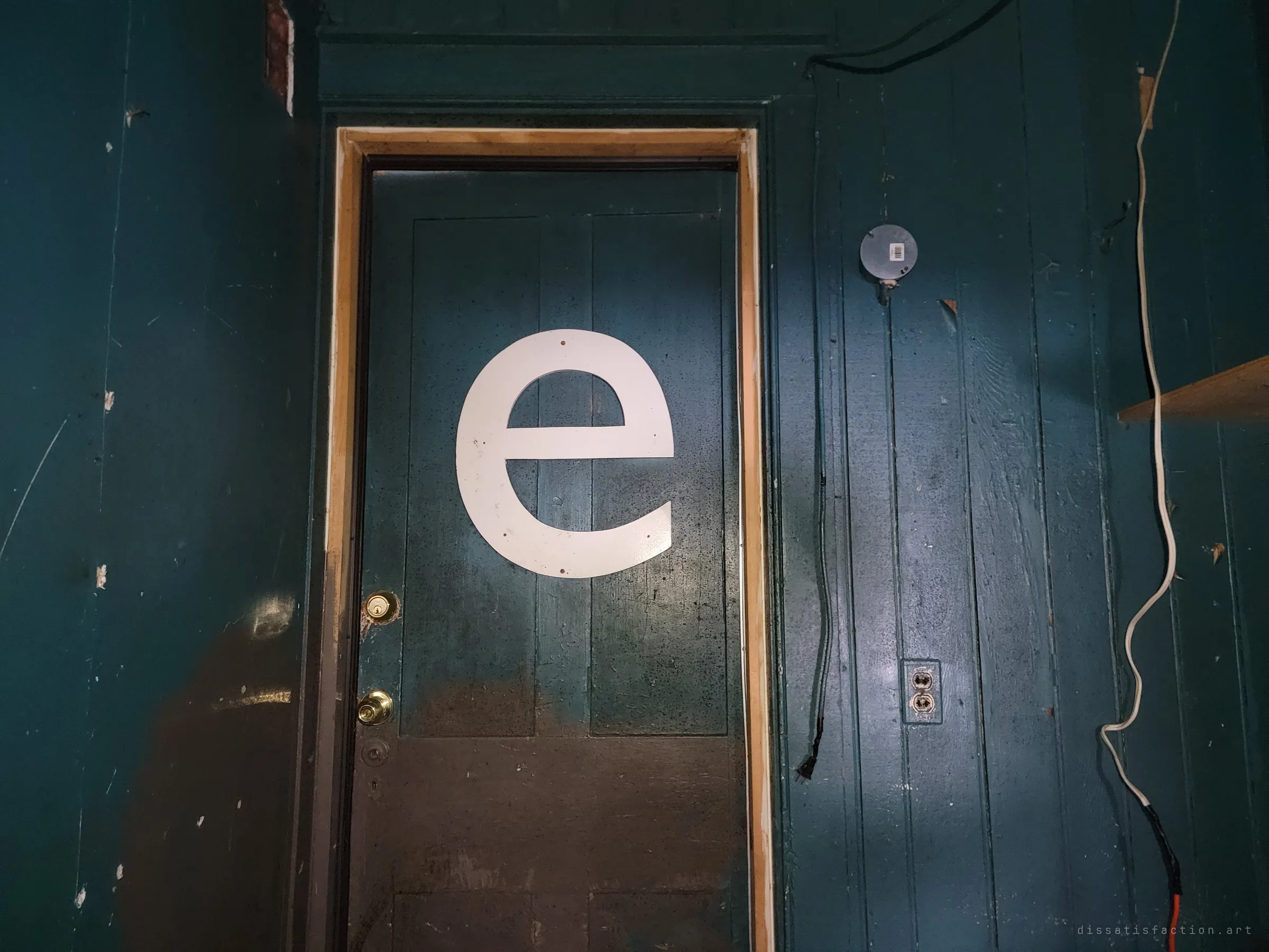 A large white lowercase letter e is fixed to a dark teal panelled door, framed in fresh raw wood against teal board walls with dangling wires. The odd salvaged sign letter is the one playful detail left in a gutted house.