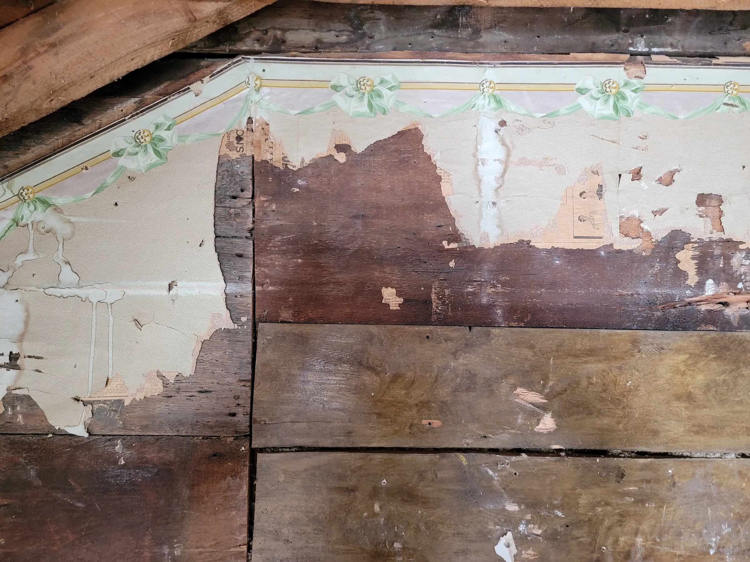 Peeling floral wallpaper with a green ribbon-and-bow border comes away from rough wooden boards under a sloped ceiling, exposing old newsprint pasted beneath. The layers show how many decades the walls have held.