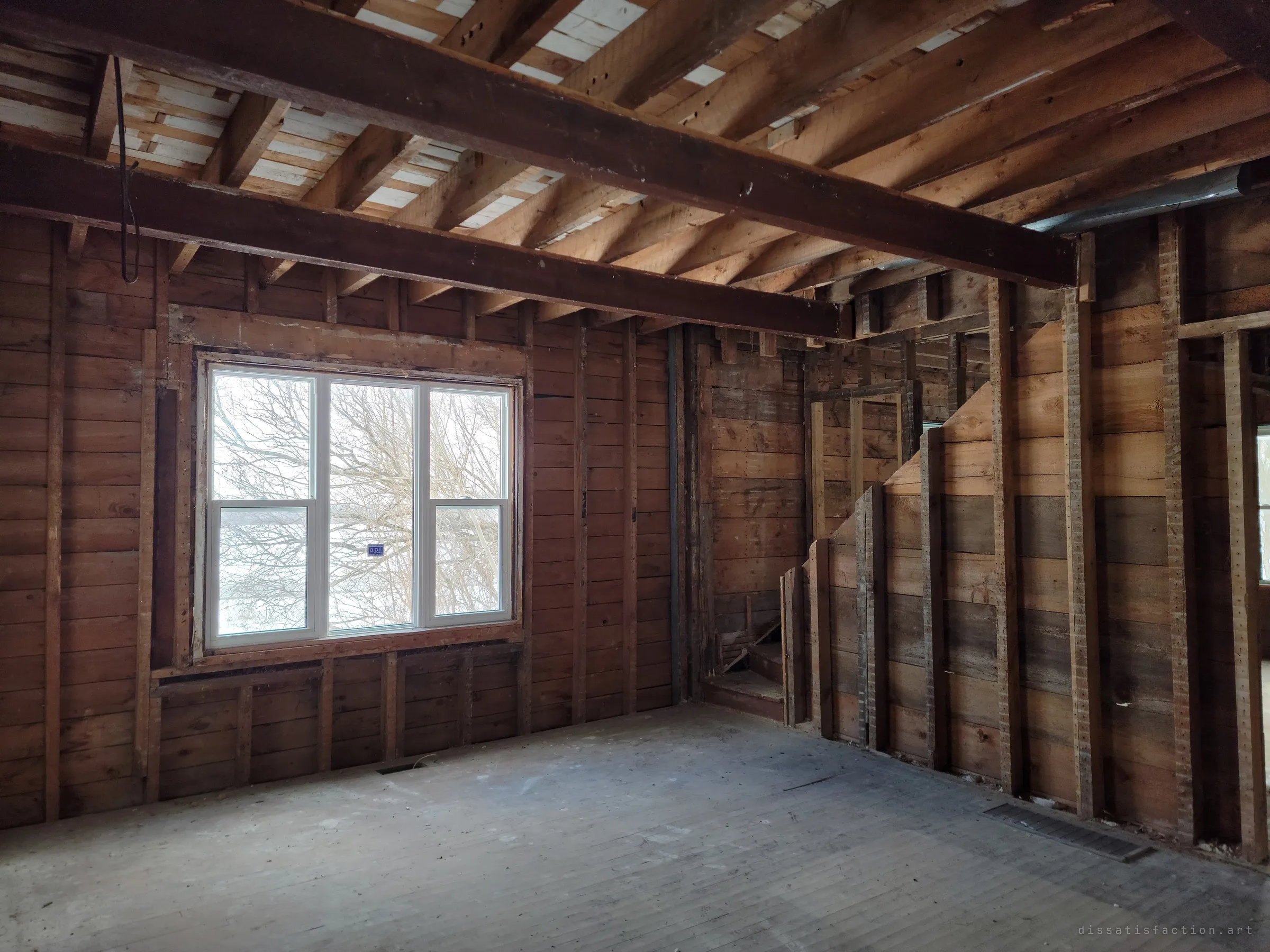 A room stripped to its wooden plank walls and exposed ceiling joists, with a new white window looking onto bare winter branches and a narrow stair boxed in at the side. Only the structure is left.
