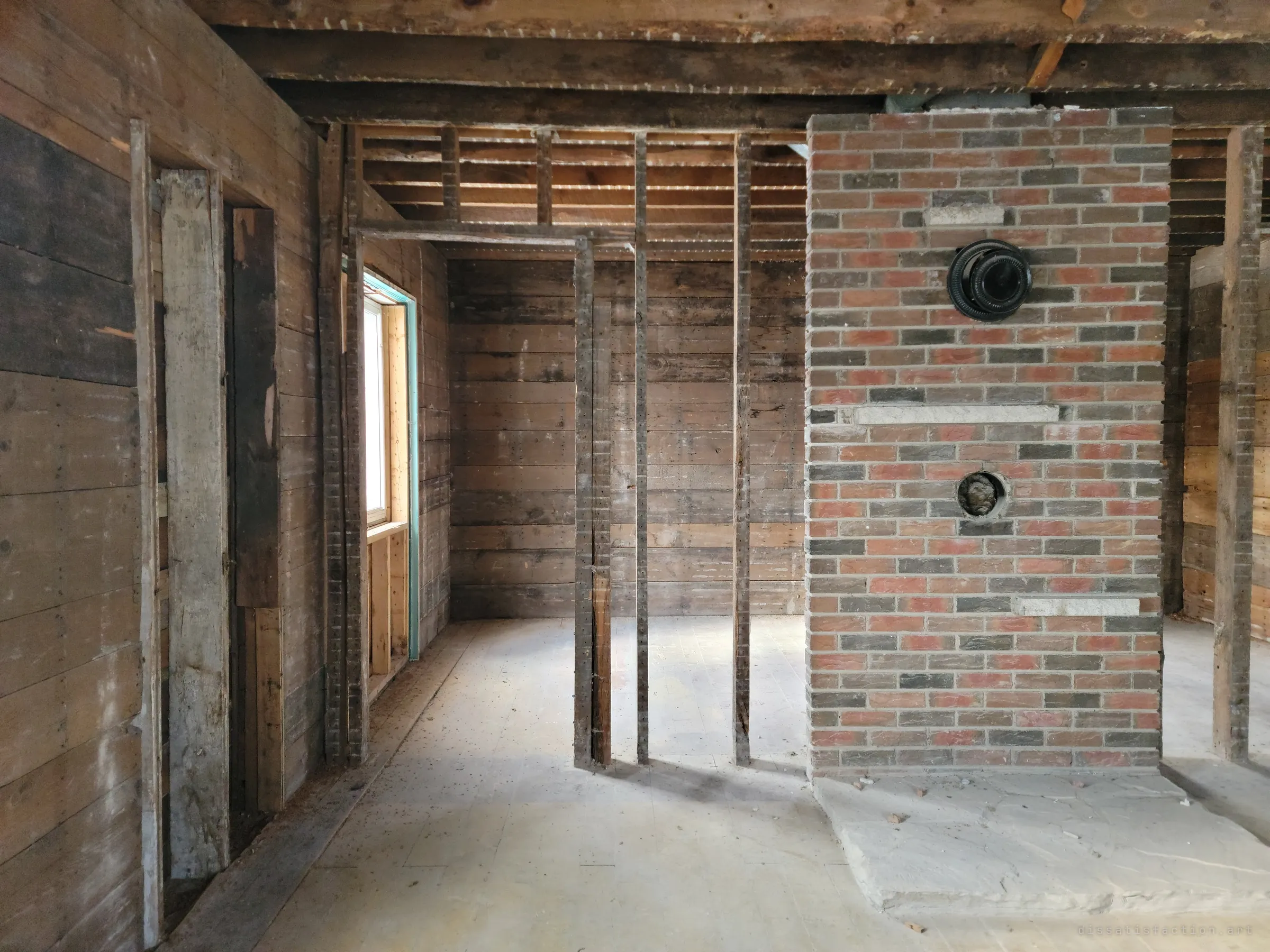 A red and grey brick chimney stands alone in a gutted ground floor, its stovepipe openings capped, surrounded by bare studs and plank walls on a dusty floor. It marks where the old stove once heated the house.