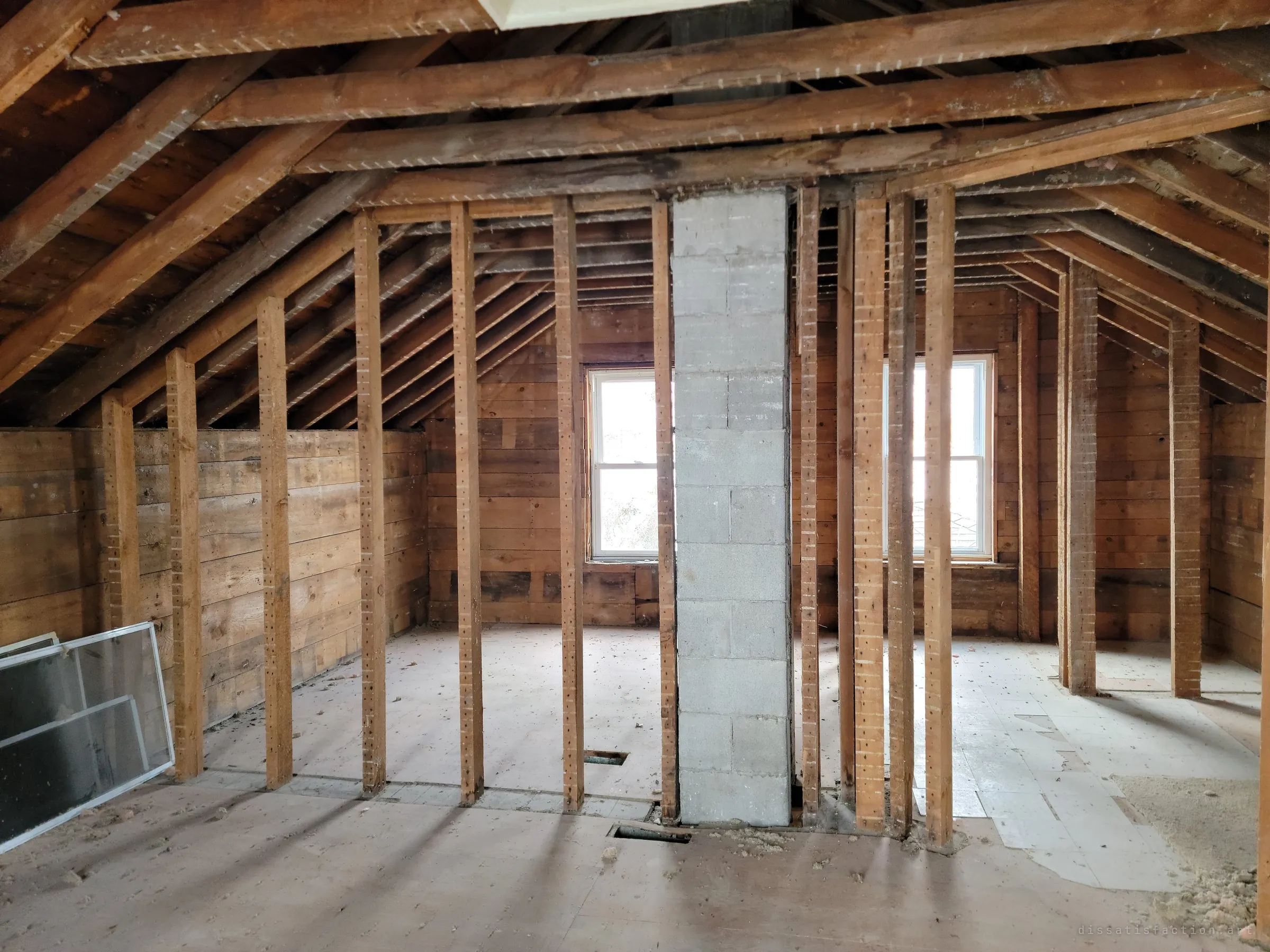 An attic under a steep timber roof, split by a row of bare studs and a grey concrete-block chimney, with two windows letting in pale light and old window screens leaning against the wall. The upper floor is stripped to its frame.