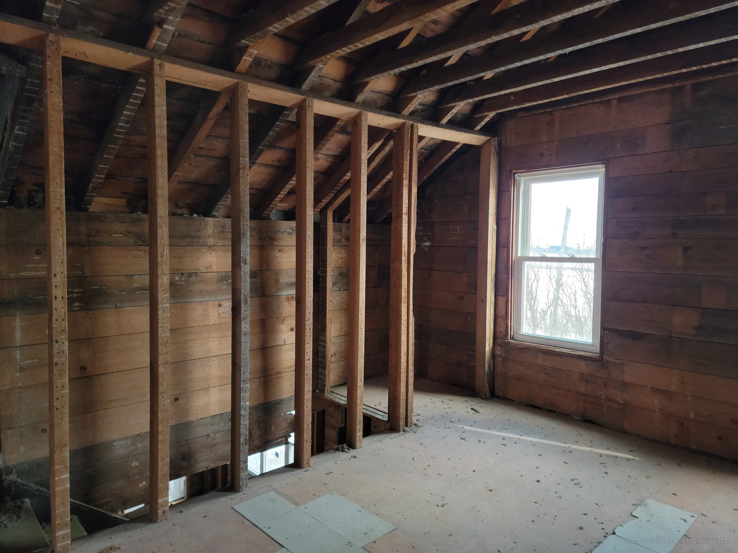 A sloped-ceiling upstairs room of rough boards and bare studs, with a window looking out over a snowy field and brush, and a gap in the floor opening onto the stairwell below. Winter light makes the empty room feel cold.