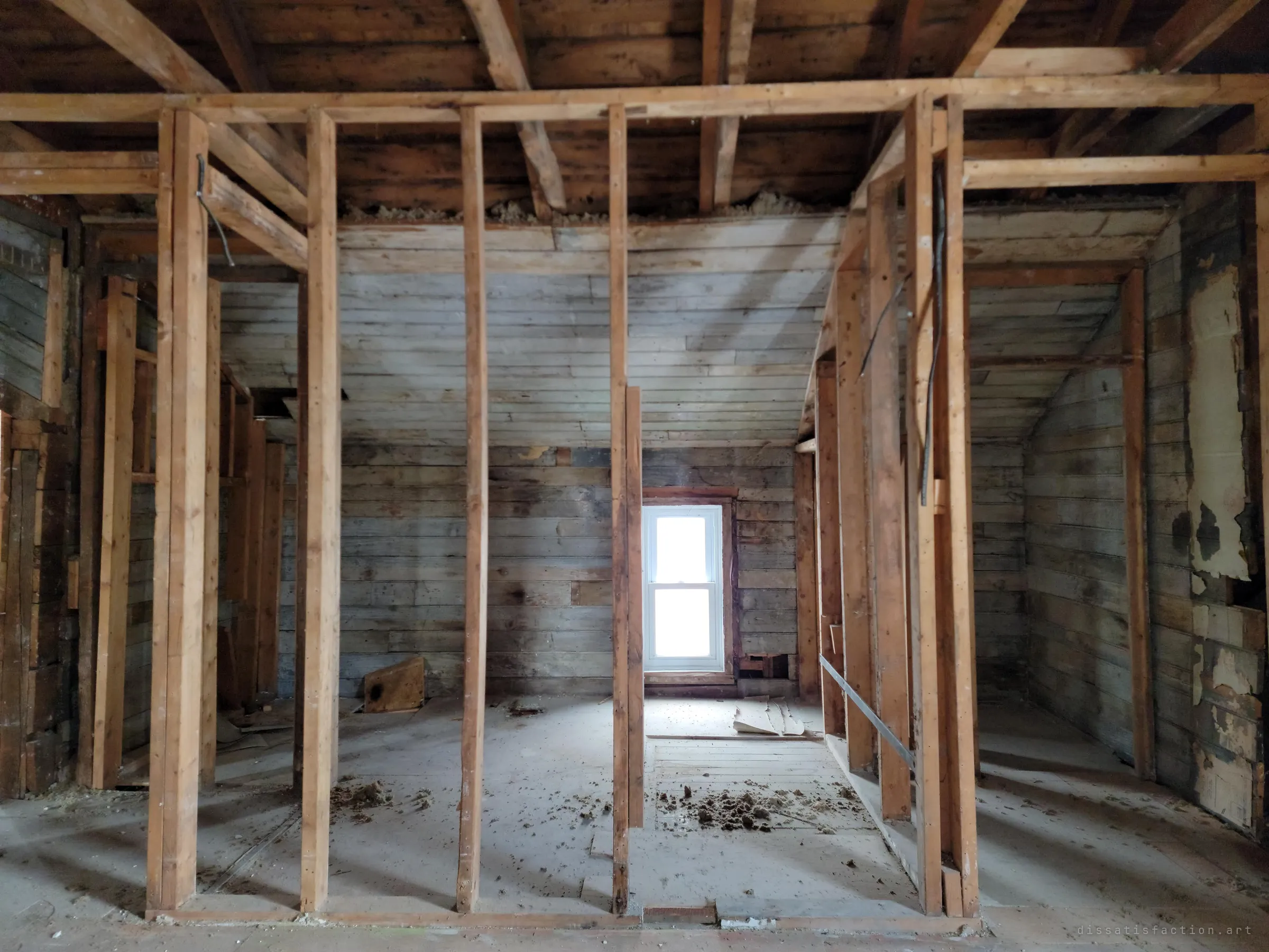 New pale stud framing marks out rooms inside a weathered grey board attic, with a small window glowing at the far end and insulation scattered on the floor. The house sits halfway between demolition and rebuilding.