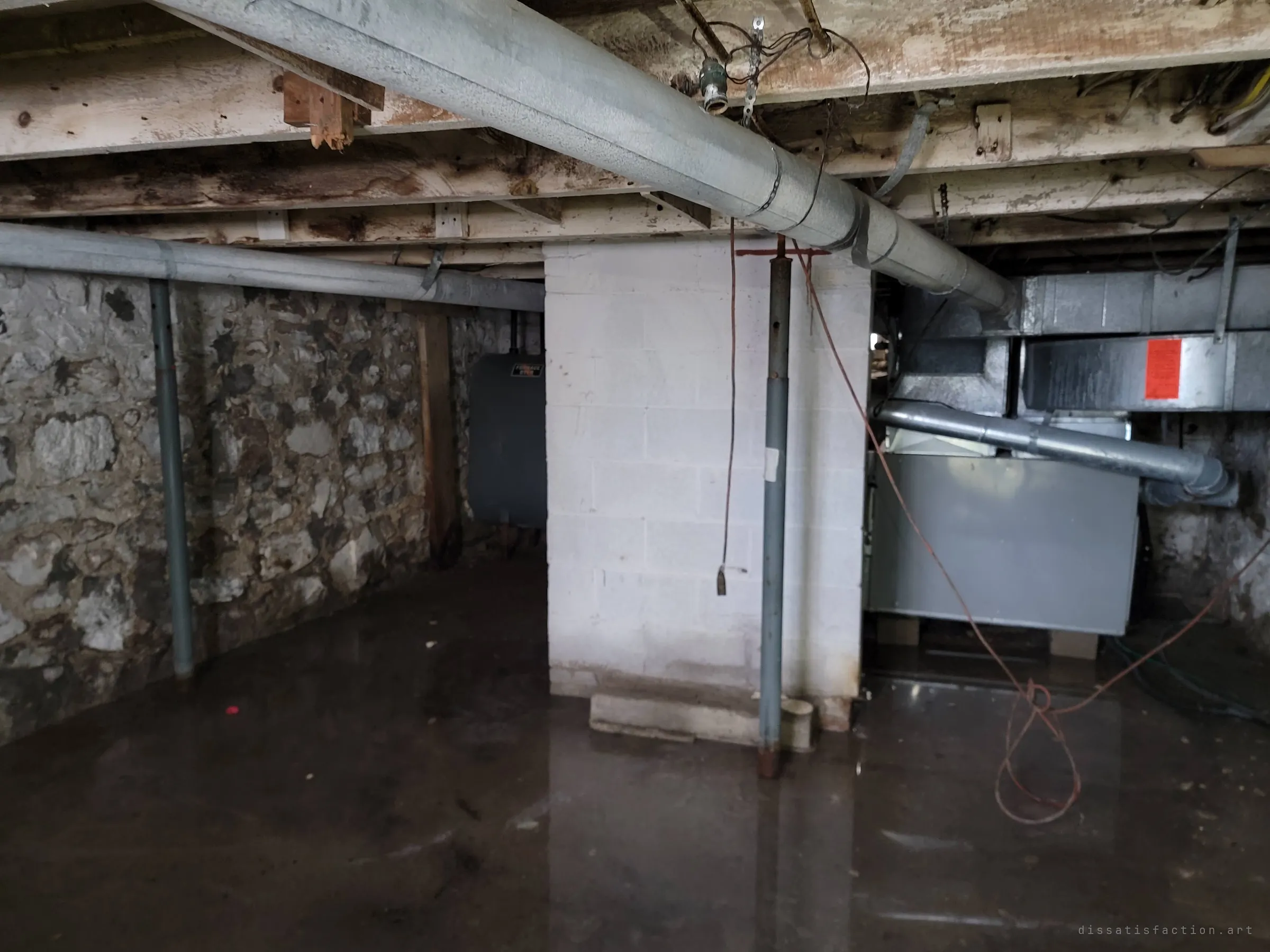 A flooded basement with a fieldstone foundation wall, a painted block pillar, steel support posts and a furnace under wrapped ductwork, standing water reflecting the ceiling. An orange tag hangs on the ductwork, its print unreadable.