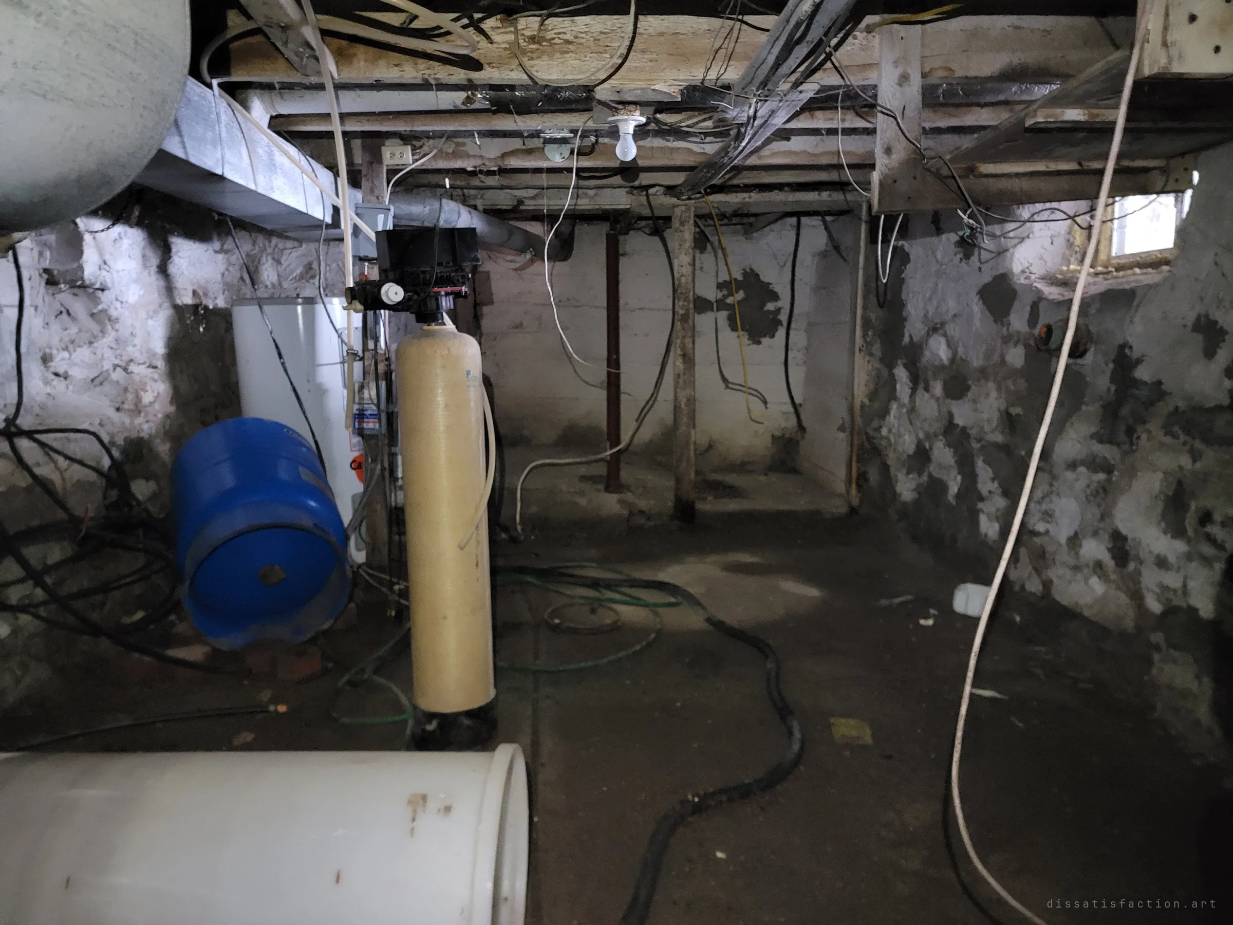 A dim stone-walled basement crowded with a water softener tank, a blue pressure tank, a water heater and tangled hoses and wires under a bare bulb. The mechanical heart of the house left behind in the damp.