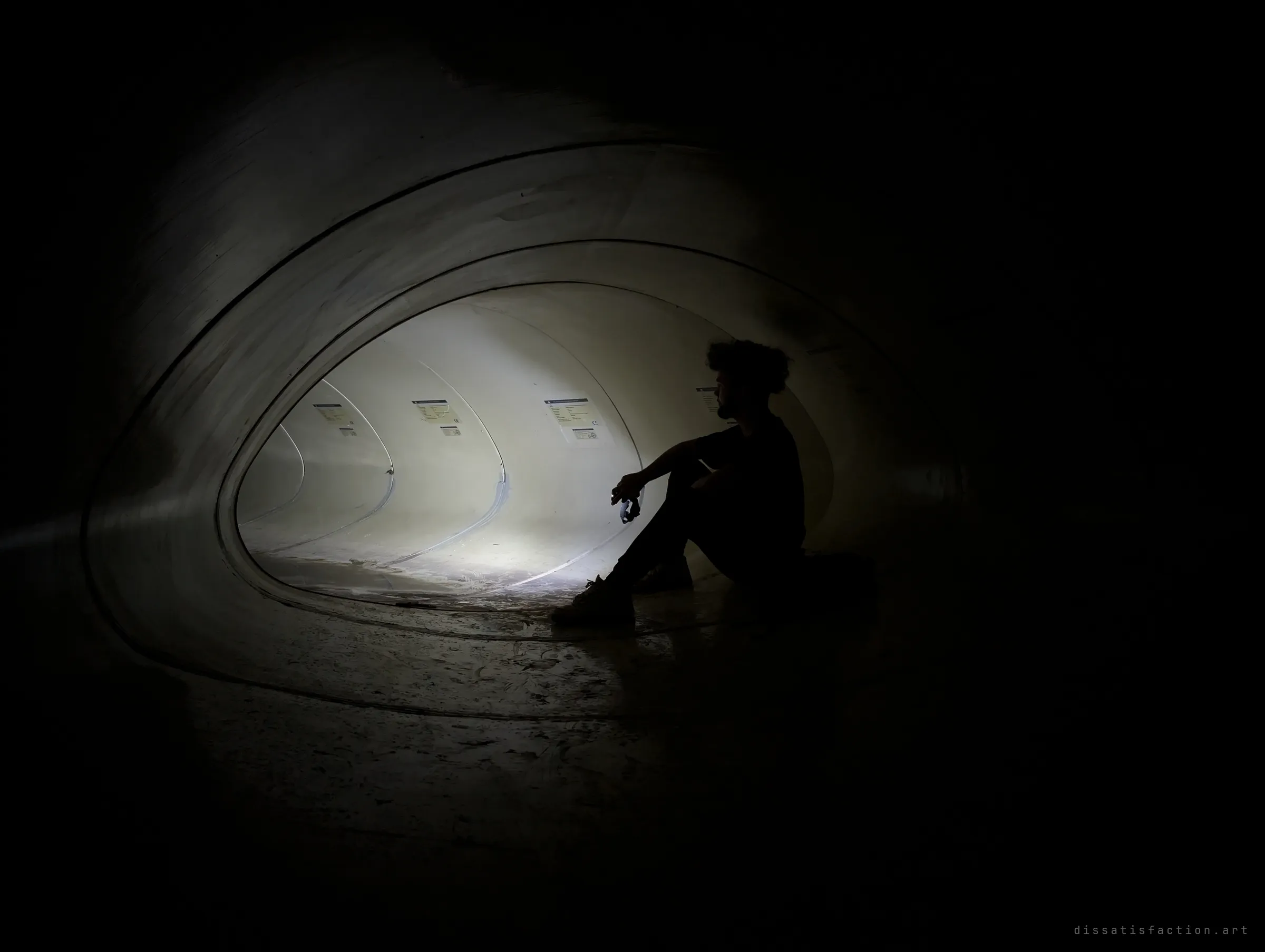 The same sitter, now nearly a silhouette against the glowing inner joints of the pipe, with darkness closing in around the frame.