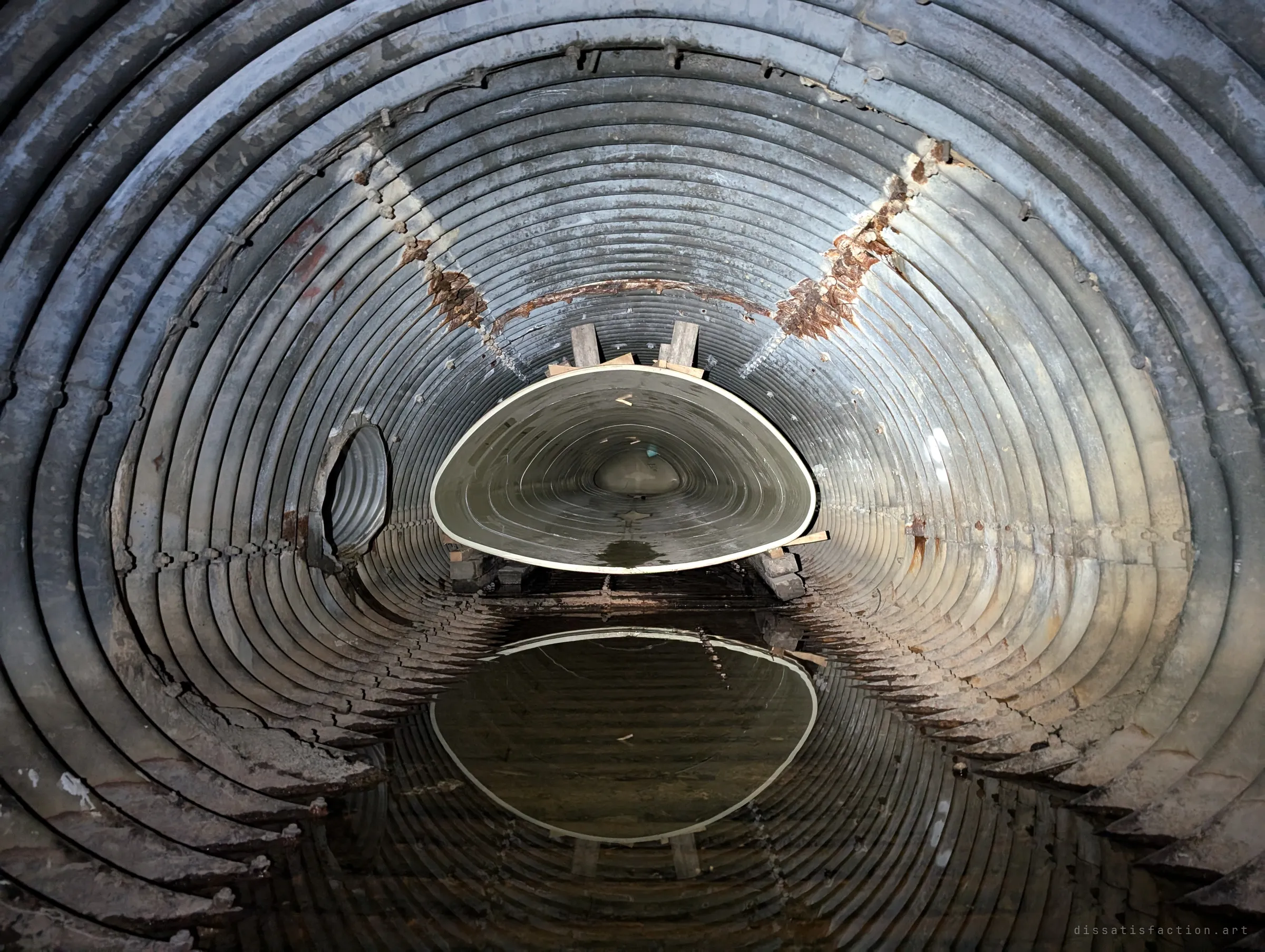 Inside the old corrugated steel culvert, rusted seams ring a pool of still water that mirrors the pale liner pipe resting on timber blocks.