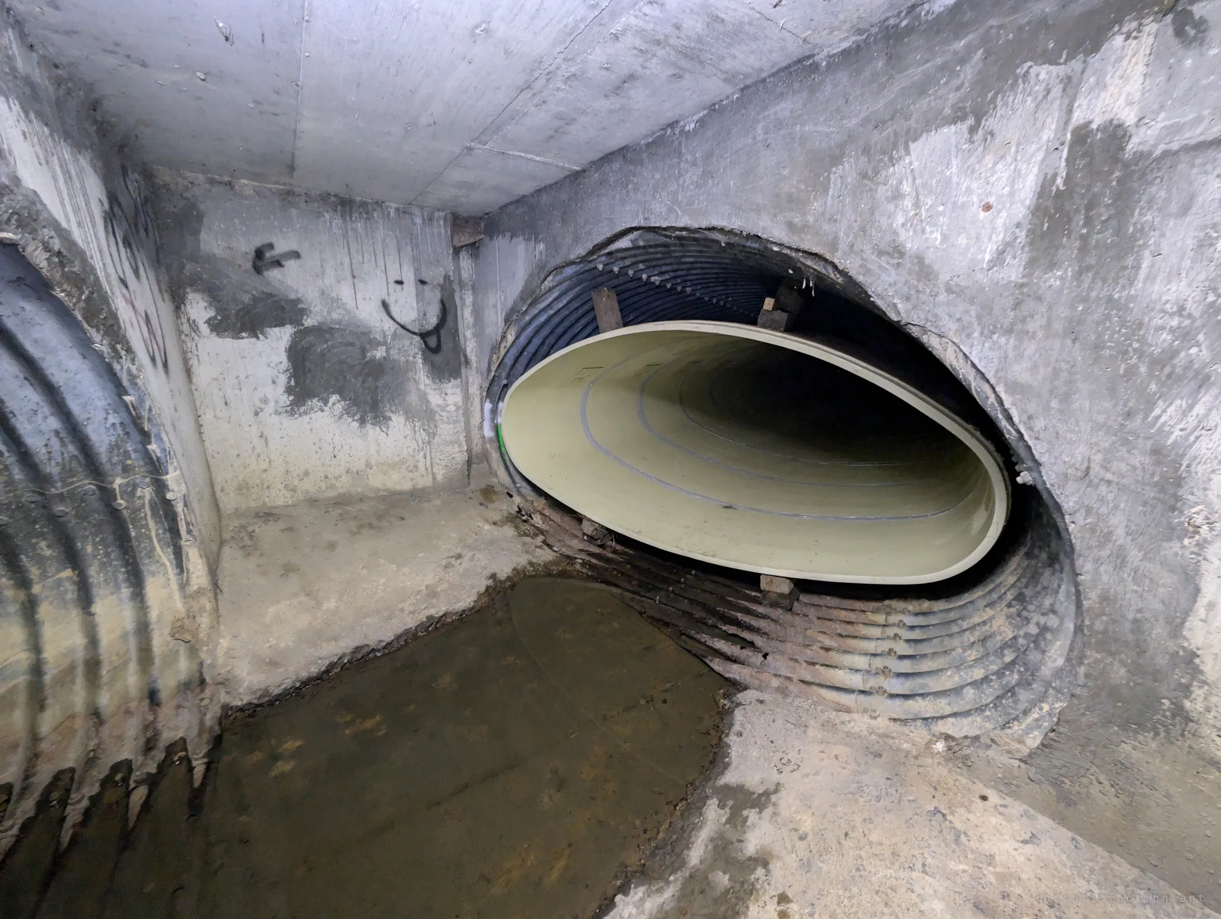 A concrete junction chamber where the liner pipe emerges from a broken-out opening in the wall, beside a corrugated side pipe and a small painted face.