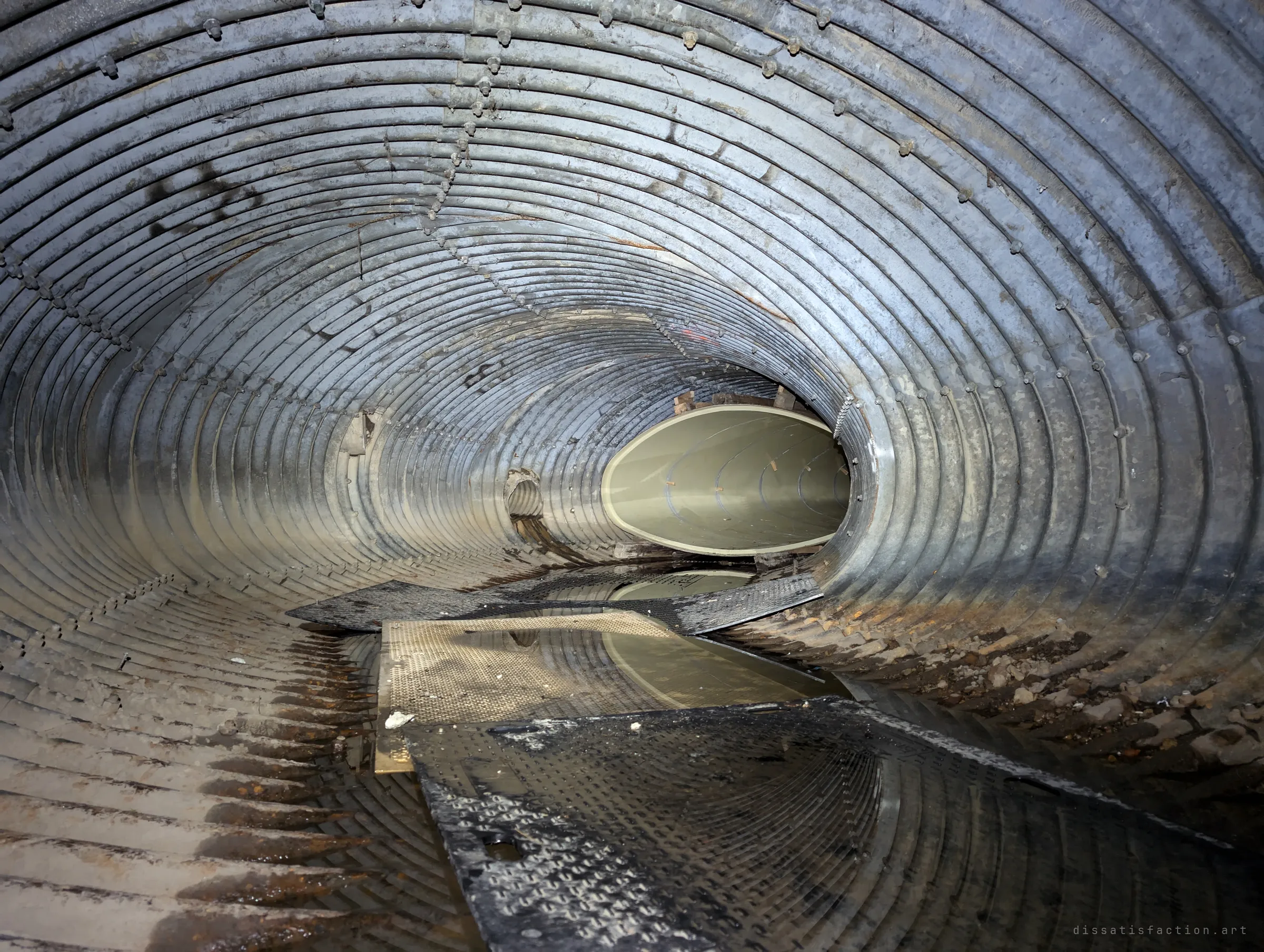 A long galvanised corrugated tunnel with loose steel plates on the wet floor, leading to the mouth of the liner pipe further in.