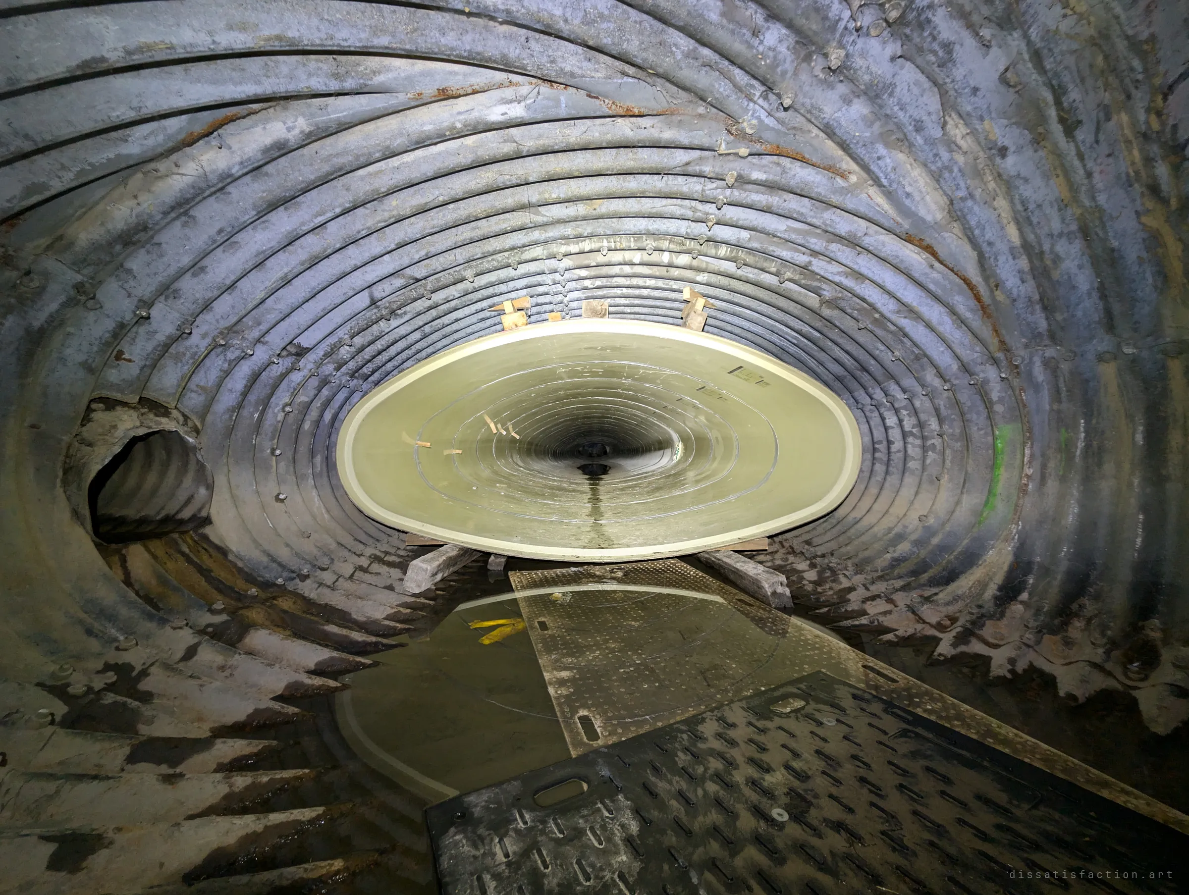 A new pale fibreglass liner pipe sits on timber blocks inside a larger rusted corrugated-steel drain, half-sunk in shallow water with rubber mats underfoot. It shows the drain mid-relining, the old pipe swallowing the new.