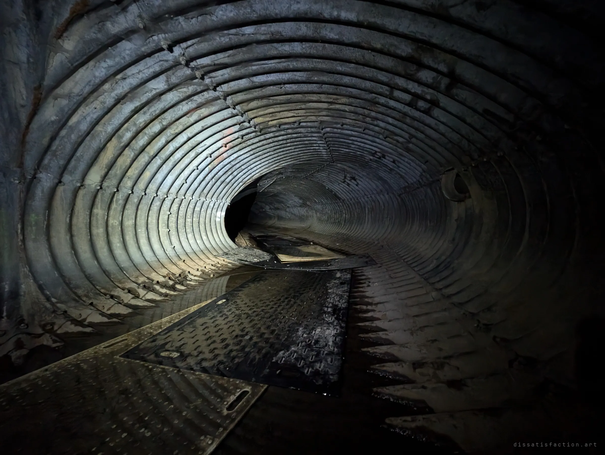 A dark corrugated-steel drain curves away to the left, lit from one side, with overlapping rubber mats laid over standing water. The ribbed walls catch the light like a coiled spring.