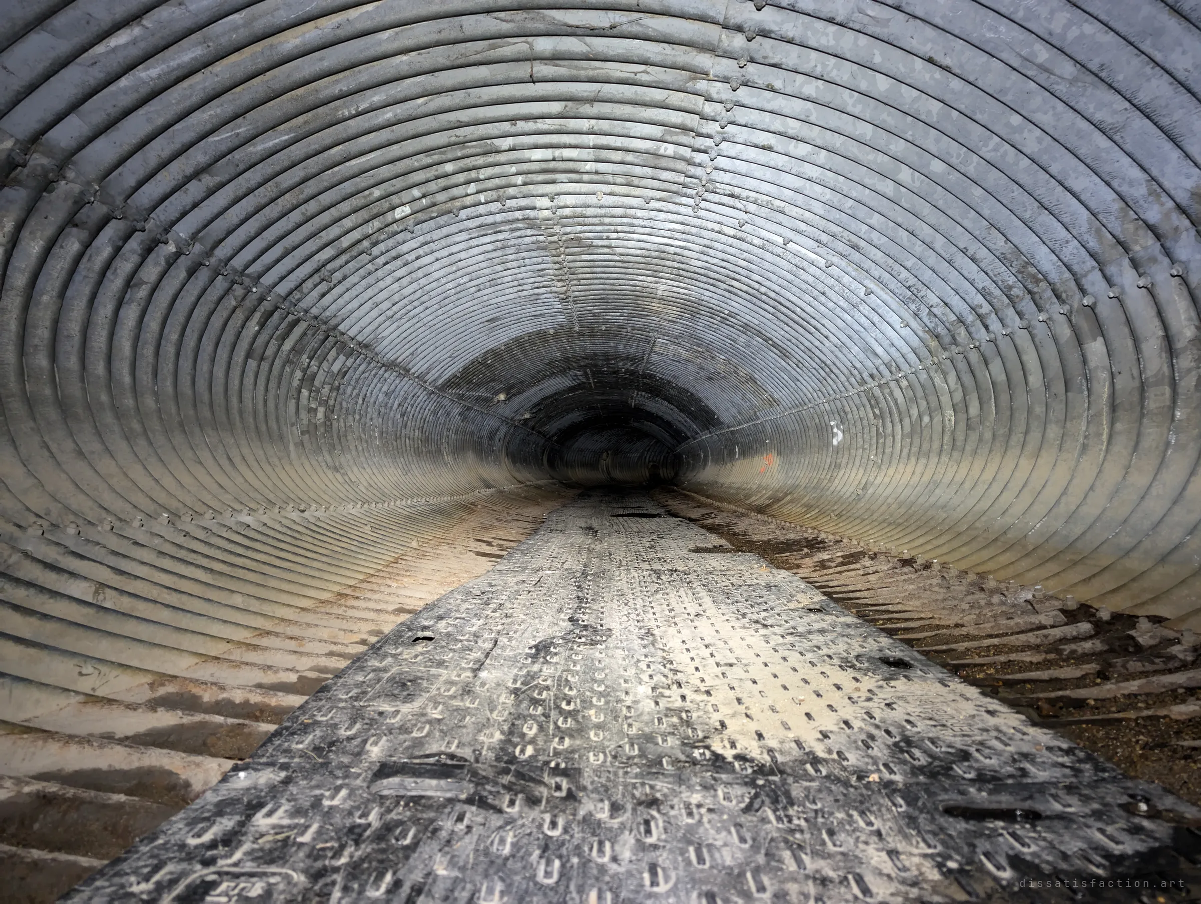 A long, perfectly straight run of galvanised corrugated pipe with a rubber mat walkway down its centre, the ribs rushing toward a dark point. The symmetry makes the tunnel feel endless.