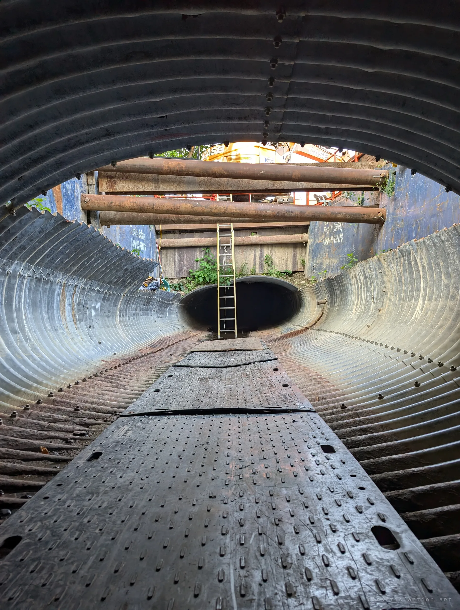 From inside the corrugated pipe, the roof opens onto a braced construction shaft where daylight, a ladder and steel struts appear before the next pipe section. It shows where the drain was cut open for the relining work.